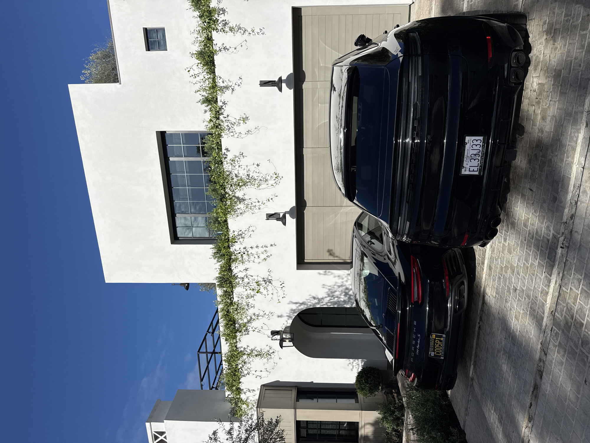 The image depicts two black Porsche vehicles parked on a patterned driveway in front of a modern, white building. A vine with white flowers grows along the wall. The building has various window styles and a garage door. The sky is clear and blue. The license plate of the top car reads "EL33J33" and of the bottom car is "C005P1", suggesting a location in California. The architecture of the building suggests a residential area within the city of Los Angeles, United States of America.