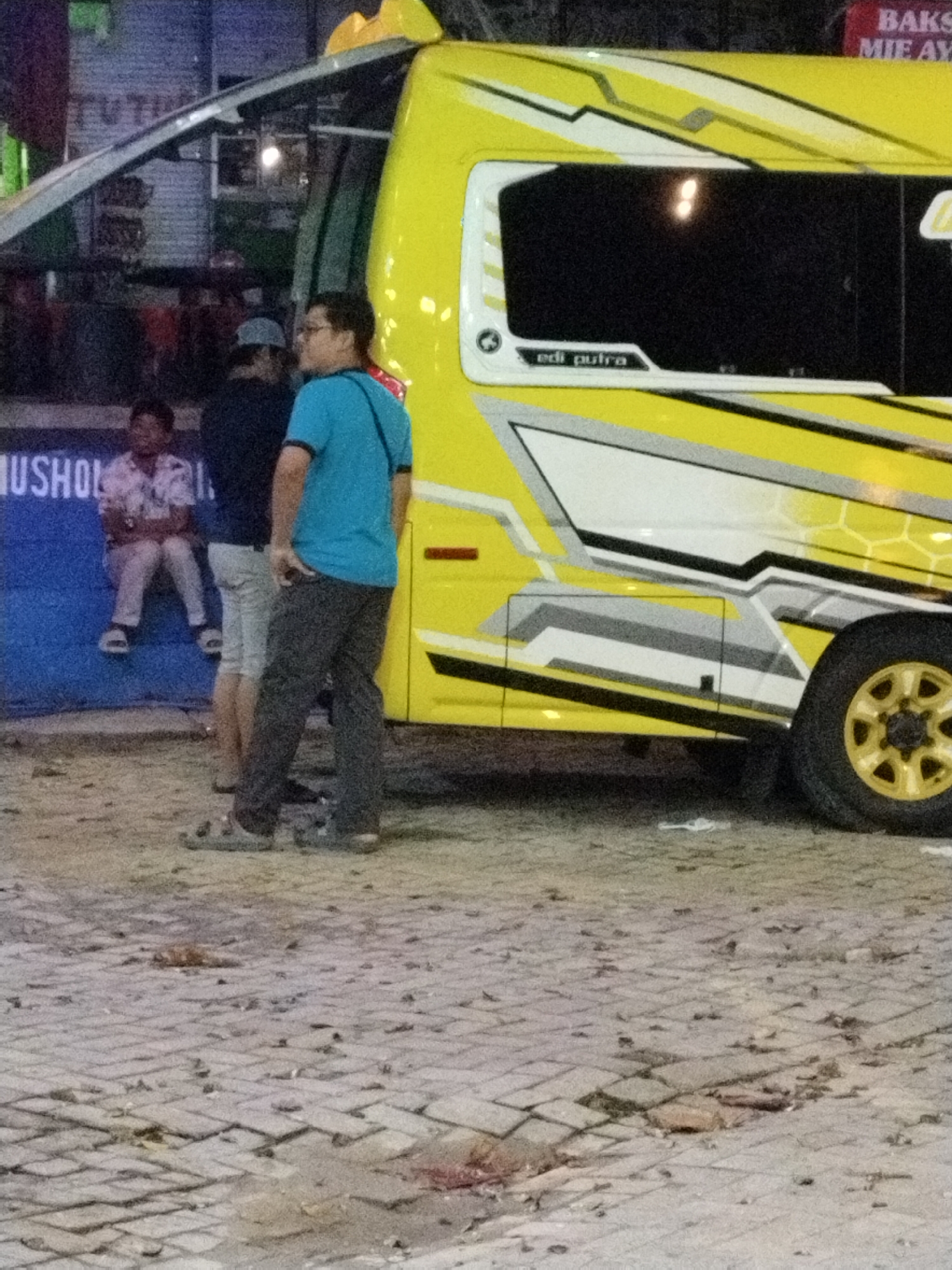 In the city of Prigi, Indonesia, a yellow truck with a white and black design is parked on a cobblestone street. Three people are standing near the truck. The person on the right is a man wearing a blue t-shirt and dark pants, facing away from the camera with his hand in his pocket. To his left, another person, possibly a man, is wearing a dark shirt and light-colored shorts. A third person, a child, is sitting on a blue wall to the left, wearing a patterned shirt and light pants. A portion of a sign that reads "USHOL" is visible on the blue wall. The background shows a building with dark windows and some red signage. The ground is covered in cobblestones with some debris and stains visible.