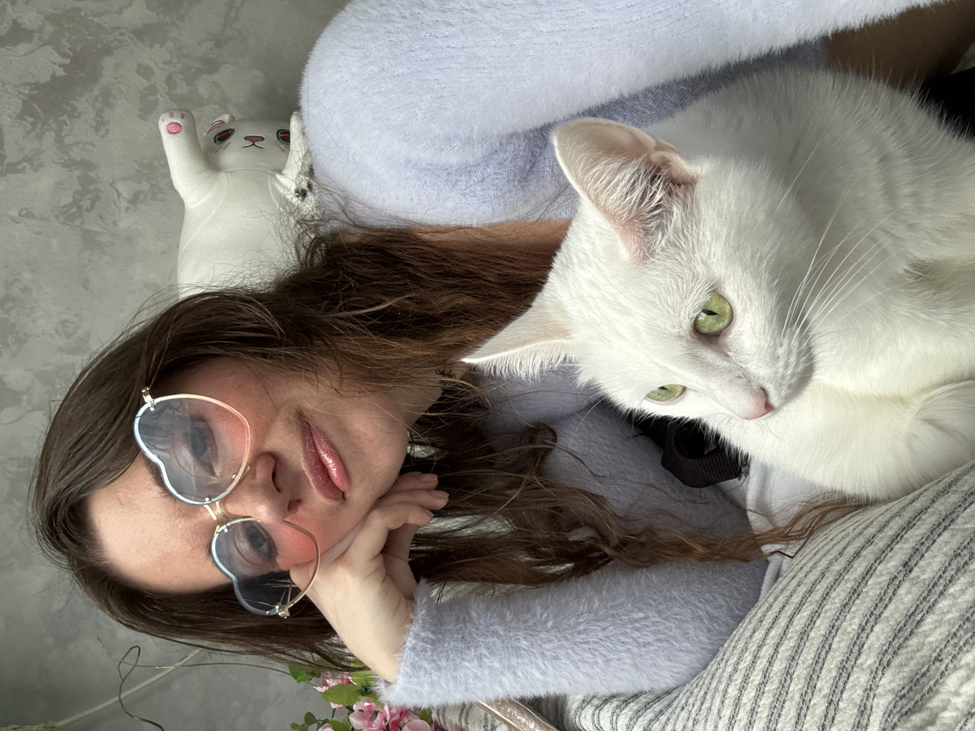 In a close-up, indoor shot, a woman with long, brown hair rests her chin on her hand, looking towards the camera. She wears heart-shaped glasses with pink and blue tinted lenses and a soft, light blue fuzzy sweater. To her right, a white cat with bright green eyes is visible, its head turned to the left, with its body mostly obscured. Behind the woman, a white stuffed cat toy is positioned upside down. The background is a textured, grey wall. There is no discernible indication of the location being in the city of Chaikovsky, Russia.