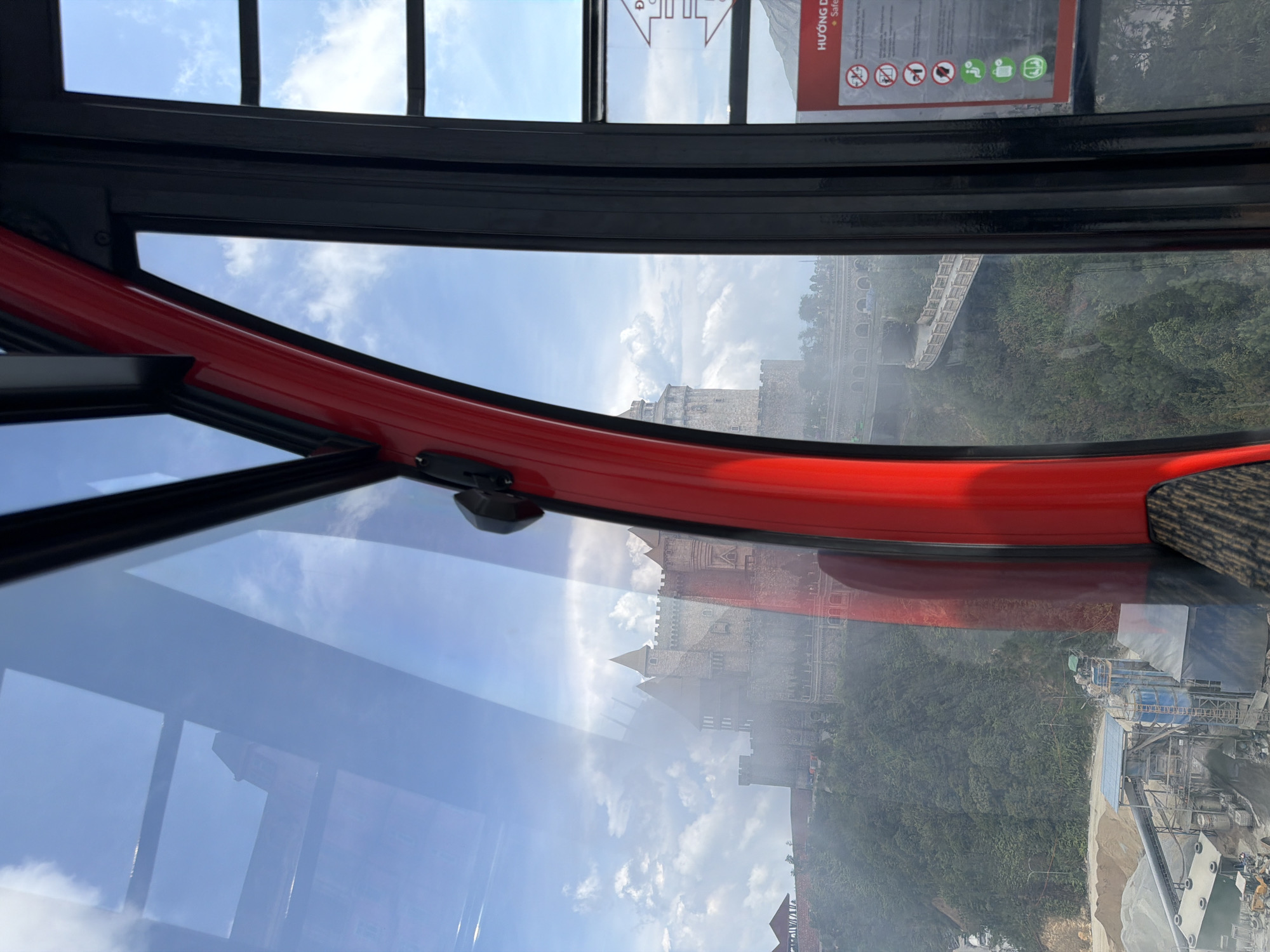 The image, taken from inside a cable car, shows a view of the Ba Na Hills in Đà Nẵng, Vietnam. The architecture visible includes a castle-like building and a long bridge. There's also evidence of construction or industrial activity in the lower right corner. The sky is visible with scattered clouds. A red interior structure of the cable car is also prominent in the foreground.