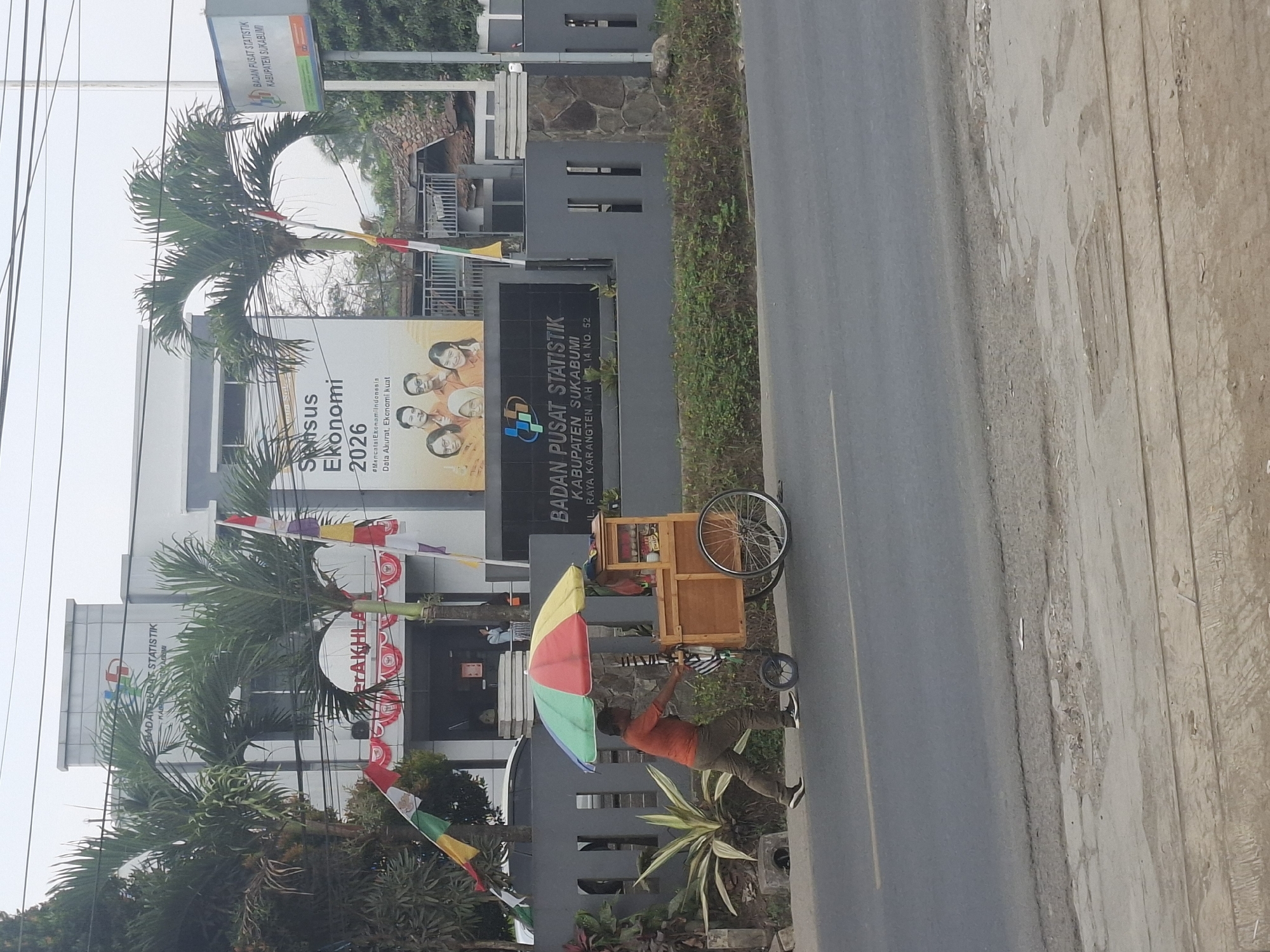 A man in an orange shirt and brown pants pushes a wooden food cart with a colorful umbrella on a sidewalk next to a road. The cart has a large wheel at the back and a smaller wheel at the front, and appears to be selling snacks. Behind the cart is a building with "BADAN PUSAT STATISTIK KABUPATEN SUKABUMI" written on it in large letters, indicating the location as the Central Statistics Agency of Sukabumi Regency. The building also features a banner with "Sensus Ekonomi 2026" and images of people. Flags are visible on poles in front of the building. The environment appears to be a moderately urban area with trees and a paved road.