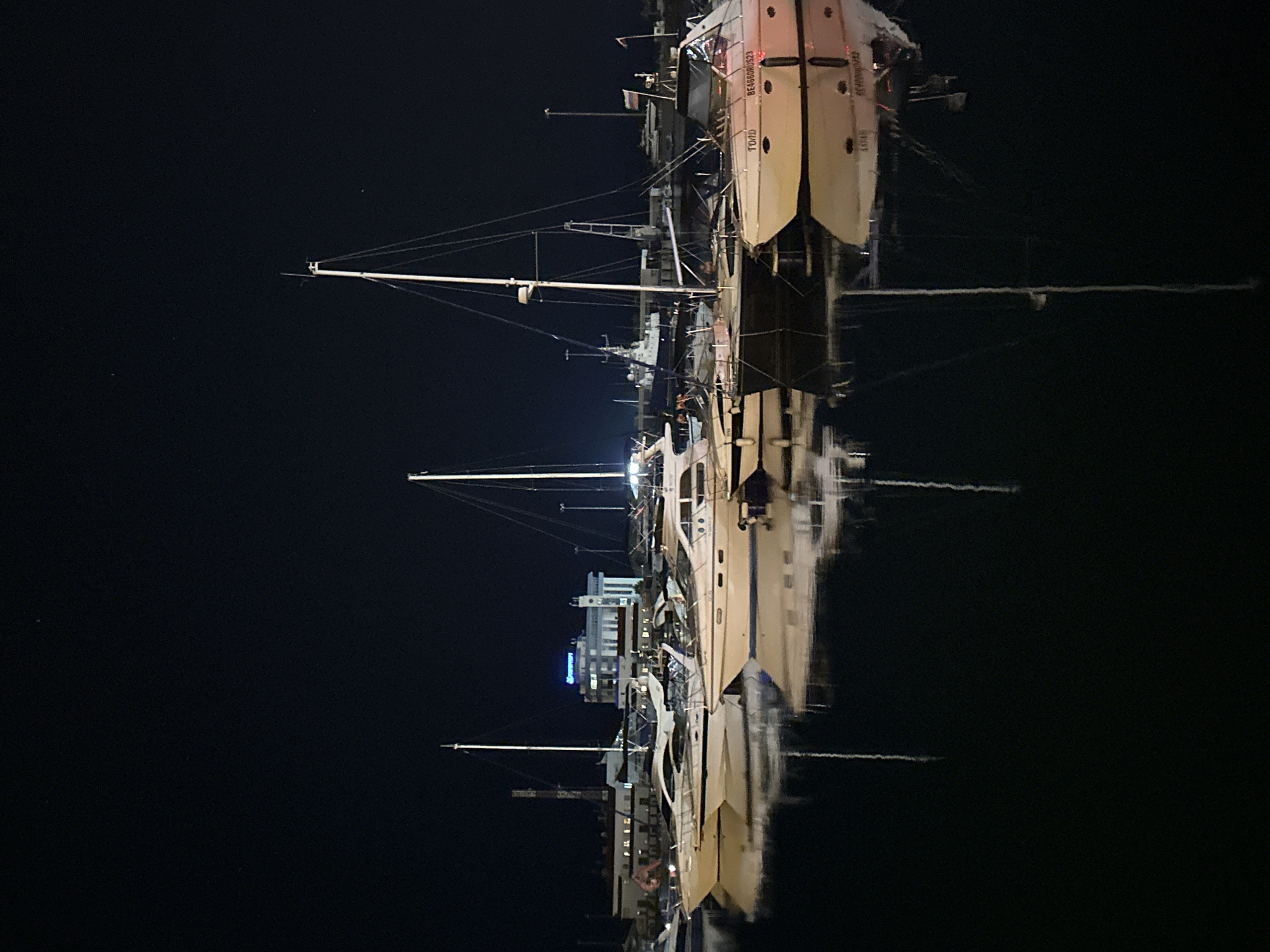 This is a nighttime photograph of a marina in Sochi, Russia, showing several sailboats docked. The image is oriented vertically, with the water and reflections appearing at the bottom. The scene is dark, indicating it's night, with lights from the boats and a building in the background illuminating parts of the scene.
The main subjects are the sailboats, with their masts and rigging clearly visible. One sailboat, larger than the others, has text on its side. Part of the text reads "GOLD" in Cyrillic script and "BE4550RUS23". The building in the background appears to be a modern structure with multiple stories and illuminated windows. The water is calm, creating clear reflections of the boats and the building.
The activity taking place is simply the boats being docked at the marina. There are no people visibly engaged in any specific activity, adding to a sense of quiet stillness of the night. The overall mood of the image is peaceful and serene, emphasizing the tranquil atmosphere of the marina at night. The location cues of Sochi, Russia, are provided as context, but are not explicitly visible in the image itself, other than the style of boats and possibly architecture of the building, which might be characteristic of the region.