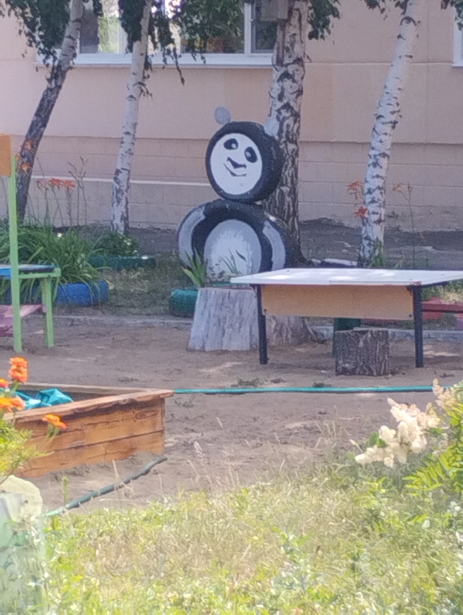 The image shows an outdoor scene that appears to be a playground or a recreational area. In the background, there is a building with light-colored walls and windows. Several birch trees are visible, with their distinctive white bark.
The central focus of the image is a decorative figure made of tires, resembling a panda. It is constructed from stacked tires painted with black and white markings to create the panda's face and body. The panda figure is placed near a tree stump.
In the foreground, there is a wooden sandbox with orange flowers growing beside it. A green hose or pipe runs across the sandy ground. To the left of the panda figure, there is a green metal table and some blue-painted tires. White flowers are visible on the right side of the image, amidst green foliage. The ground is primarily sand and dirt, consistent with an outdoor play area.
The location is identified as the city of Kostanay, Kazakhstan. The scene suggests a public space, possibly a kindergarten or a park, set up for children's activities. The overall atmosphere is bright and appears to be a daytime scene.
