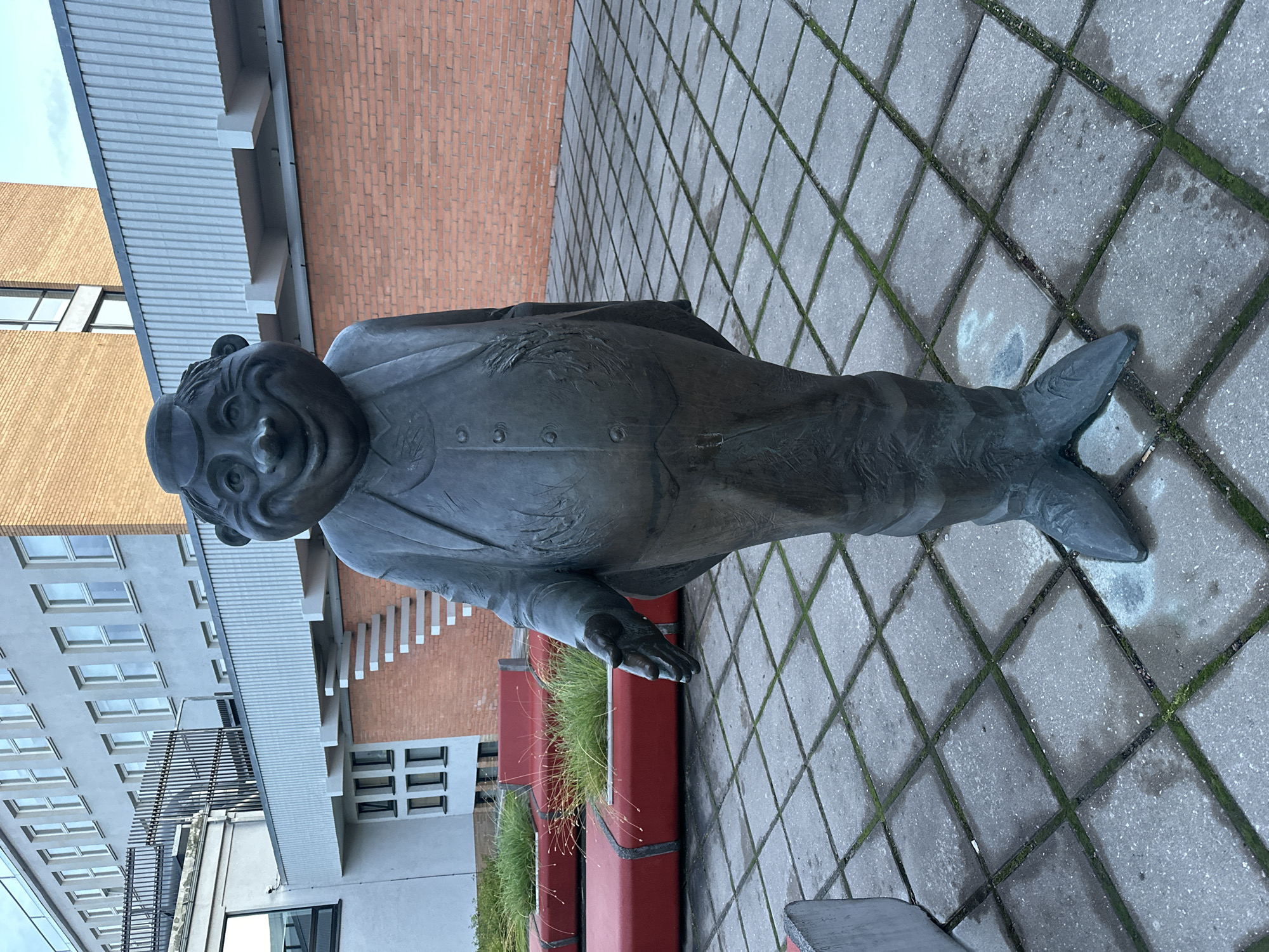 A bronze statue of a jovial, portly man stands in an outdoor paved courtyard. The statue is of a man wearing a suit with buttons down the front and a cap. His left arm is extended outward, as if offering a handshake. In the background, a modern building with light grey siding and orange brick accents is visible. Red planters filled with green grass are to the left of the statue. The courtyard is paved with grey square tiles, some of which have patches of moss. The statue is located in Tallinn, Estonia.