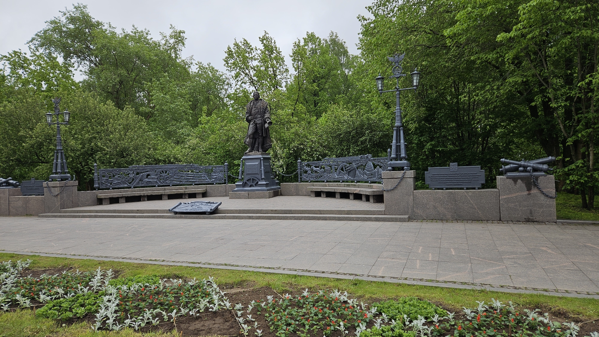 The image shows a monument in a park, possibly a memorial dedicated to Peter the Great based on the statue. The scene includes a statue of a man in period clothing, standing on a pedestal, likely the central figure of the monument. Flanking the statue are decorative benches with detailed metalwork along the back, possibly depicting scenes related to naval history or military achievements.
Additional elements of the monument include cannons displayed on raised platforms and ornate street lamps that add to the memorial atmosphere. The entire setup is on a paved area, surrounded by greenery, including trees and flower beds in the foreground. The overall impression is that of a historical landmark or memorial site in a public park. The city is likely St. Petersburg, Russia, given the statue's resemblance to Peter the Great and the city's history, but this is not certain from the image alone.