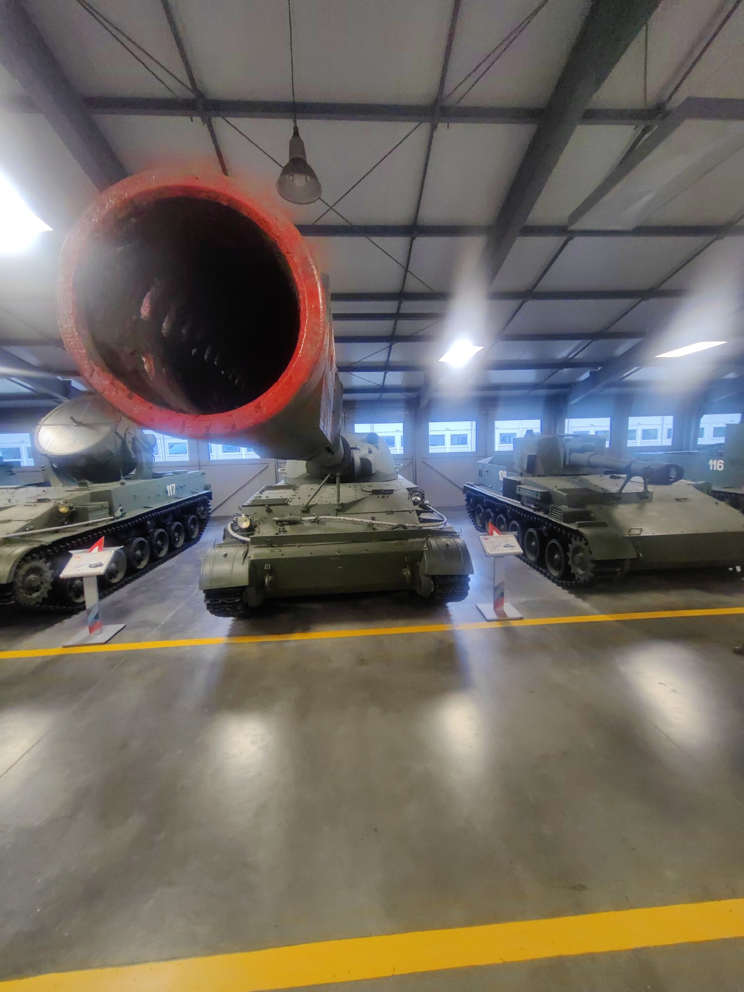In the city of Kubinka, Russia, within the Central Armed Forces Museum, two self-propelled artillery pieces are displayed on a concrete floor marked with yellow lines. The larger of the two, a self-propelled howitzer, is positioned in the foreground, its barrel extending upwards and painted red at the muzzle. The second artillery piece, smaller and to the left, is also green and displays the number "117" on its hull. A third, partially visible artillery piece is located to the right. The indoor environment features a high ceiling with exposed metal beams and white paneling, illuminated by overhead lights. Several windows are visible in the background, offering a view of the exterior. No people are present in the image.