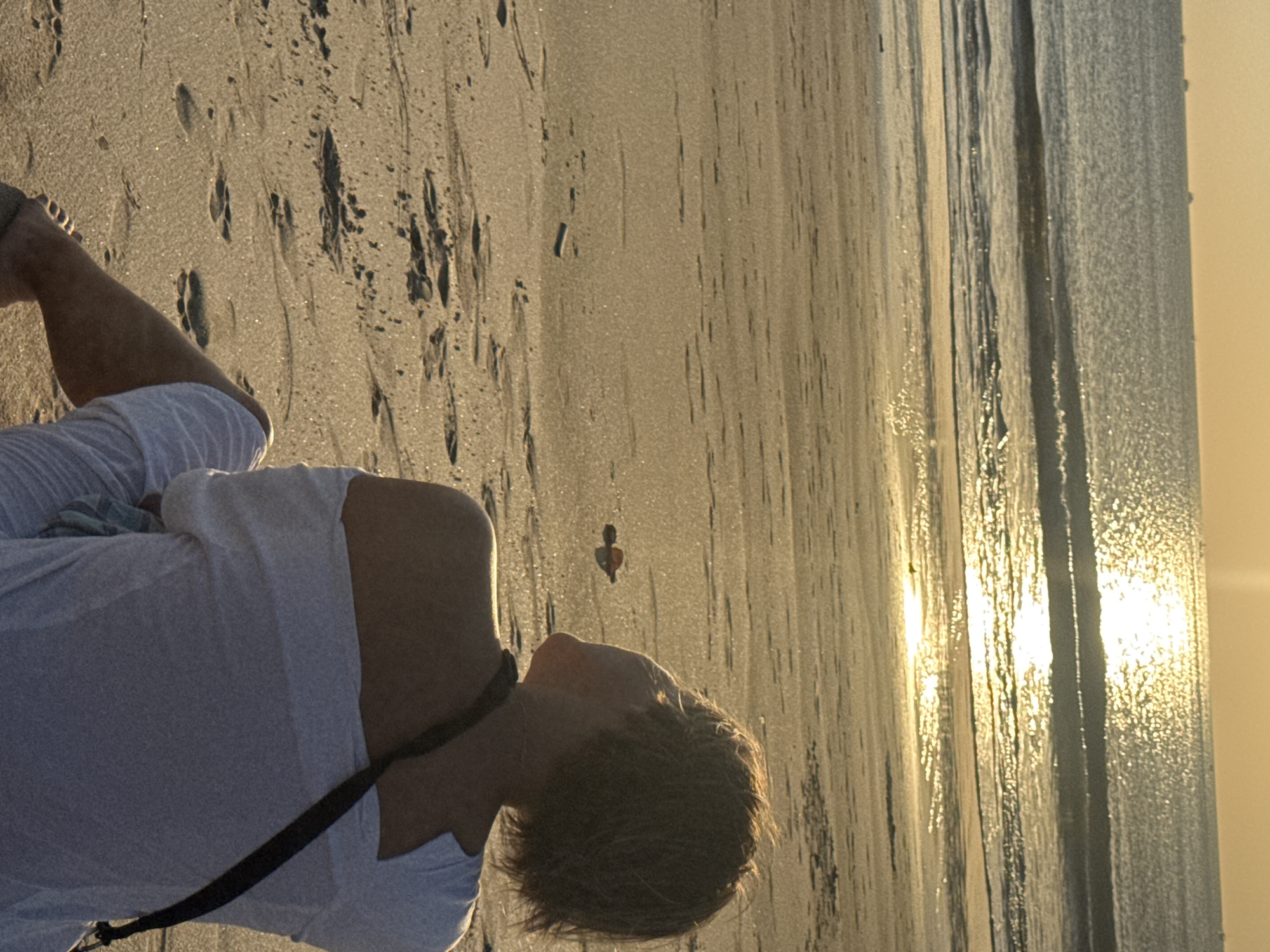 The image depicts a solitary individual sitting on a sandy beach, facing the ocean. The person, viewed from behind and the side, has short, light brown hair and is wearing a white off-the-shoulder top. Their body is angled towards the sea, and they appear to be looking upwards towards the horizon. The beach is marked with footprints in the wet sand, suggesting recent activity. The ocean stretches out to the horizon, with gentle waves rolling in. The sun is low in the sky, casting a warm, golden light that reflects brightly on the water's surface and the wet sand. The overall atmosphere is serene and tranquil, with the setting sun creating a peaceful, almost ethereal glow. The location is specified as Kuta, Indonesia, known for its beautiful beaches and vibrant sunsets.