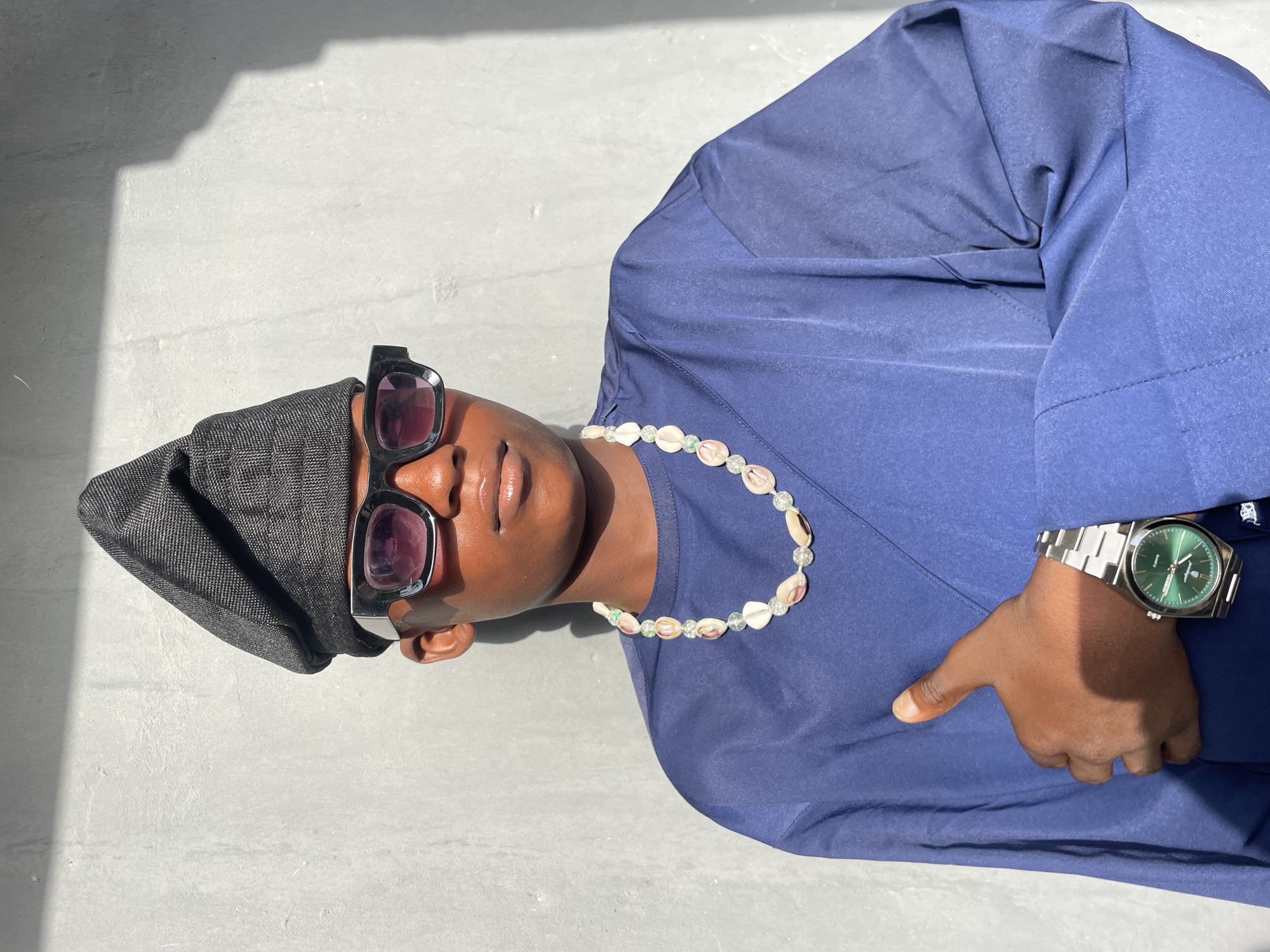 A person with a dark skin tone is captured in a top-down, slightly angled view, lying on a light grey, possibly concrete or tiled, outdoor surface. Their head is oriented towards the left of the frame, and their body extends diagonally across the image.
The individual is dressed in a dark blue long-sleeved shirt or tunic and a dark grey or black cap or head covering. They wear stylish black-framed sunglasses with dark, seemingly reddish-tinted lenses. A notable accessory is a necklace made of small, light-colored cowrie shells interspersed with clear or light green beads. On their left wrist, a silver-toned watch with a vibrant green face is visible. The watch face clearly shows the text "QUARTZ" and a small emblem, while additional fine print below the hands is illegible. The person's left hand is partially visible, resting with the thumb slightly outstretched and other fingers curled.
The scene is brightly illuminated by direct sunlight, casting distinct, sharp shadows, particularly a large angular shadow extending from the upper left, suggesting an overhead structure or building. This indicates a clear, sunny day, likely during mid-day or early afternoon. The overall atmosphere appears relaxed and casual. The setting is in Badagry, Nigeria.