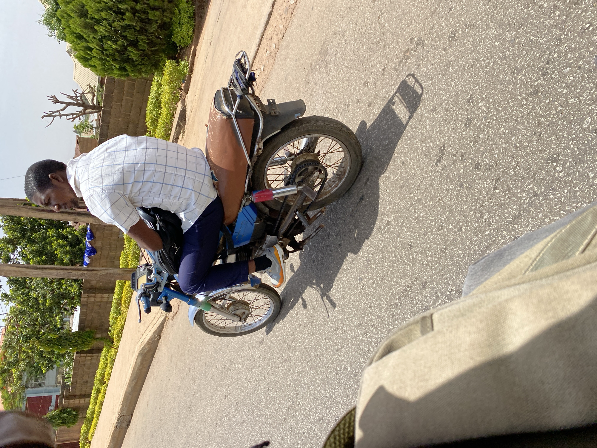 A man is sitting on a blue motorcycle on a paved road. He is wearing a white, checkered, short-sleeved shirt and dark pants with grey and orange sneakers. The motorcycle appears to be older, with a brown seat. The road is bordered by green foliage and trees on one side, with buildings visible in the background. The picture appears to have been taken in Bukuru, Nigeria.
