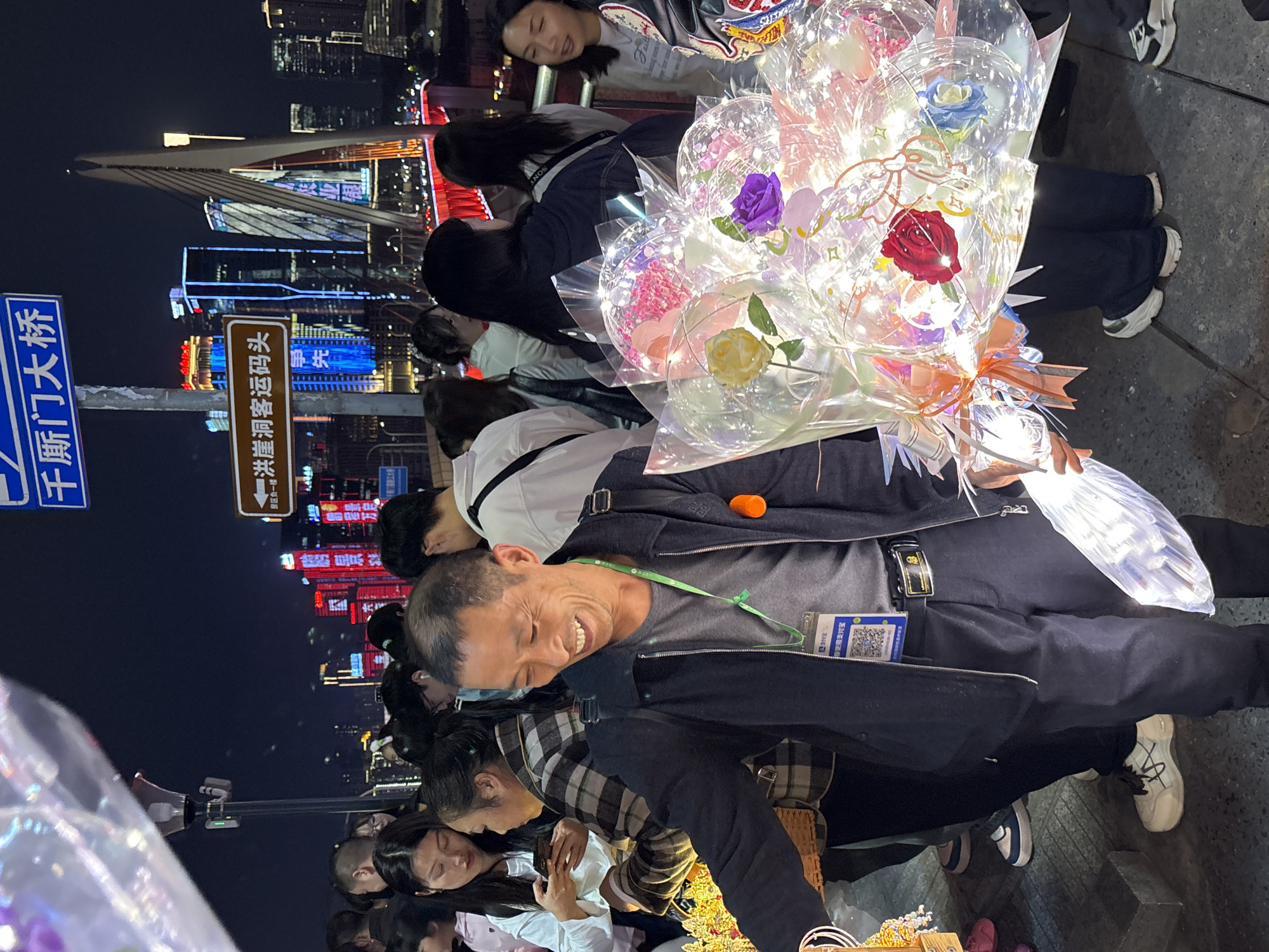 Here is a detailed description of the image:
**General Overview:**
The image captures a nighttime scene in what appears to be a bustling urban area. It features people on a sidewalk with city lights and structures visible in the background.
**People:**
There are several people in the frame. In the foreground, a man is visible, holding a bouquet of light-up floral balloons. The man is smiling. Surrounding him are numerous other people, some of whom seem to be engaging with him or the merchandise.
**Objects:**
1. Floral Balloons: Large bouquets made of flowers encapsulated in transparent balloons, illuminated with lights. The flowers come in various colors like red, pink, yellow and purple.
2. Signage: There are two signs visible. One has Chinese characters on it, "千厮门大桥" (Qiānsīmén Dàqiáo) and the other one has Chinese characters on it, "洪崖洞客运码头" (Hóngyádòng Kèyùn Mǎtou).
3. Cityscape: The background includes an illuminated bridge and tall buildings, suggesting a dense urban environment.
4. Other items: Some people carry bags and wear jackets. There appears to be a cart or stand where the flowers are displayed.
**Actions:**
The man is selling floral balloons. Other people are walking around, observing, or potentially purchasing items.
**Location:**
Based on the signage mentioning "Qiānsīmén Dàqiáo" and "Hóngyádòng Kèyùn Mǎtou", the location is likely in the Yuzhong District, Chongqing, China, near the Qiānsīmén Bridge and the Hongyadong tourist area. These are known landmarks in the Yuzhong district.
**Environment:**
The scene is set at night, with artificial lights illuminating the area. The background cityscape features high-rise buildings with many lights on, indicating a vibrant urban setting.
**Summary:**
The photo portrays a lively nighttime scene in Yuzhong District, China, with a man selling illuminated flower balloons near popular landmarks like the Qiānsīmén Bridge and Hongyadong, attracting people to the area.