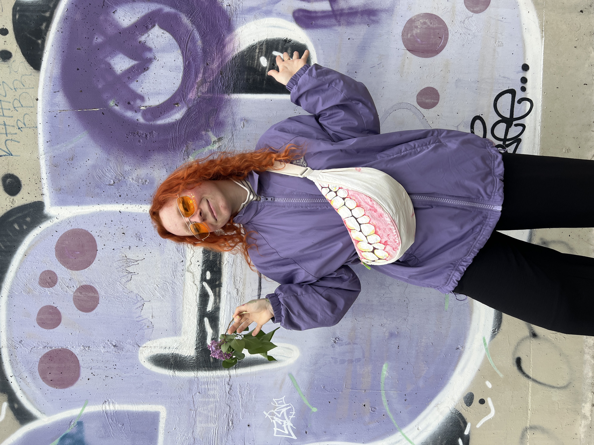 A young individual with vibrant, curly red hair is depicted lying on their back against a concrete wall covered in graffiti. They are wearing a purple lightweight windbreaker jacket, black pants, and distinctive round orange-tinted sunglasses. A white fanny pack, uniquely designed with a pattern resembling a set of human teeth and gums, is worn across their chest. The person looks towards the camera with a subtle smile, holding a small sprig of purple lilac flowers in their right hand, while their left arm is raised.
The scene is an outdoor urban setting, characterized by a concrete wall heavily adorned with layered graffiti. The predominant color of the graffiti is purple, featuring large abstract shapes, circles, and various tags in shades of purple, white, and black. Visible graffiti elements include stylized letters and numbers such as "HHS", "BB" or "QB", "22", "80", "13", along with other illegible symbols and squiggles.
The image appears to be taken during the daytime under bright, natural light. The purple attire and graffiti create a cohesive color palette with the person's red hair and the lilac flowers, conveying a relaxed and playful mood. This scene is located in Belgrade, Serbia.