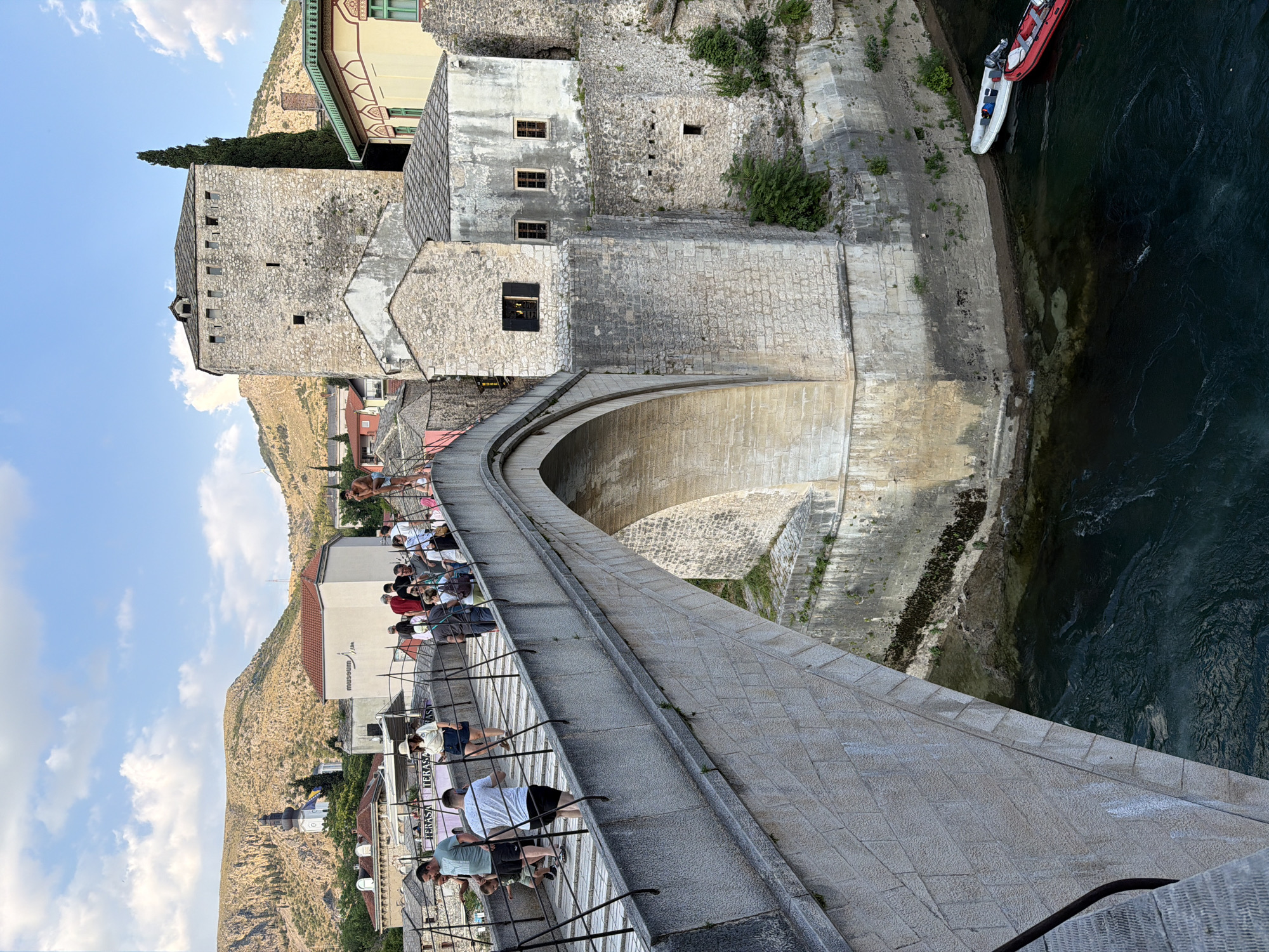 The image depicts the Stari Most (Old Bridge) in Mostar, Bosnia and Herzegovina. The stone bridge, an iconic landmark, arches over the Neretva River, which flows with a dark, turbulent current. A diverse group of tourists are crossing the bridge, some walking, others pausing. The bridge's stone construction is visible, with a cobbled pathway. On the right side of the bridge, a historic stone tower and adjacent buildings stand, showcasing old architecture with stone and stucco facades. Lush green vegetation grows along the riverbanks and on the stone structures. Two inflatable boats, one white and one red, are docked along the river's edge below the bridge. In the background, hills with sparse vegetation rise under a partly cloudy sky, and a church steeple is visible in the distance. The overall scene captures a busy, sunny day at a historic European location.