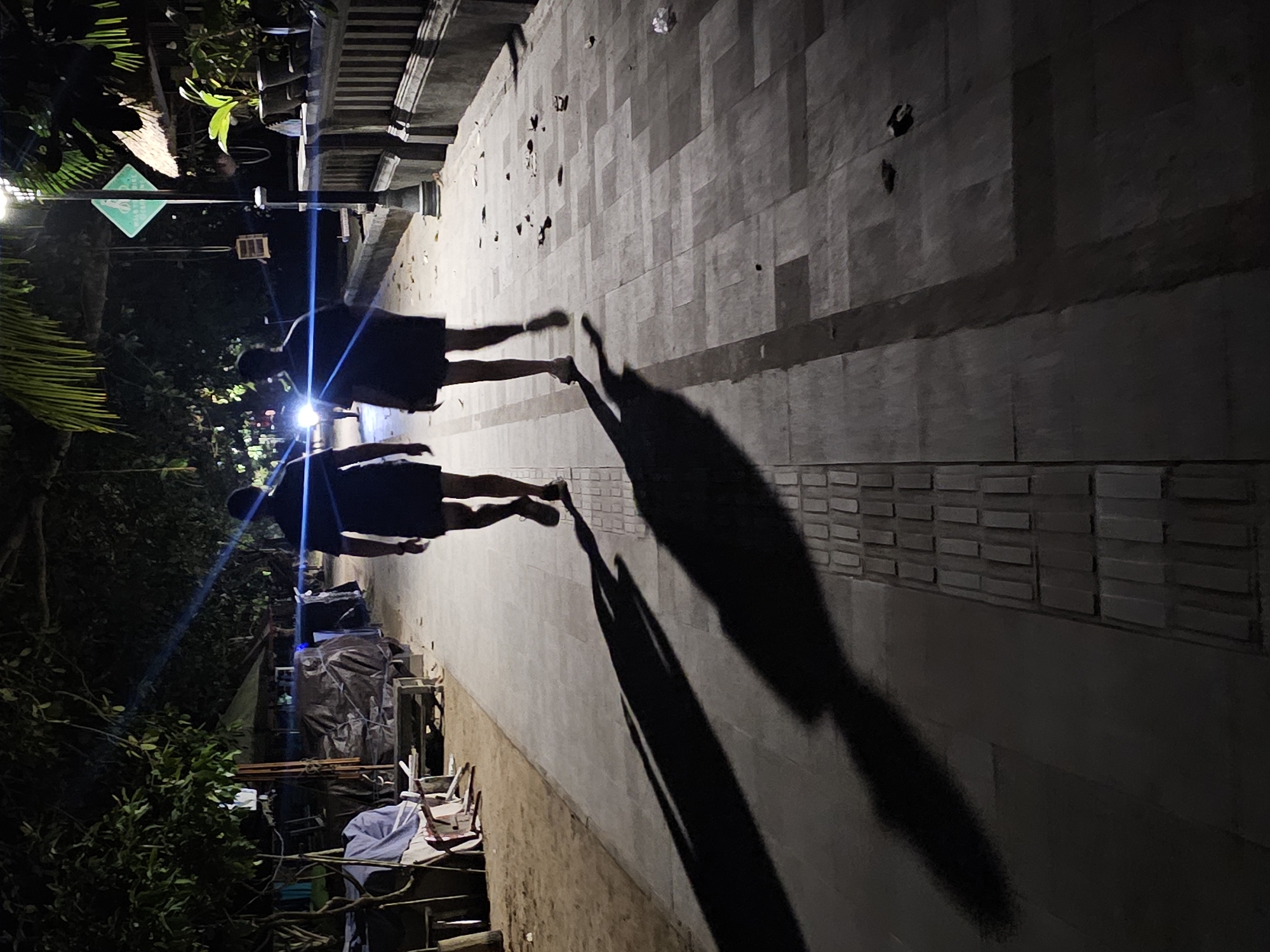 At night in Denpasar, Indonesia, two people are walking along a tiled walkway. They are wearing dark shorts and t-shirts. Their shadows are long and distinct on the walkway. The area adjacent to the walkway appears cluttered with various objects. There is a lamppost on the left side, surrounded by foliage. A green directional sign is visible near the lamppost.