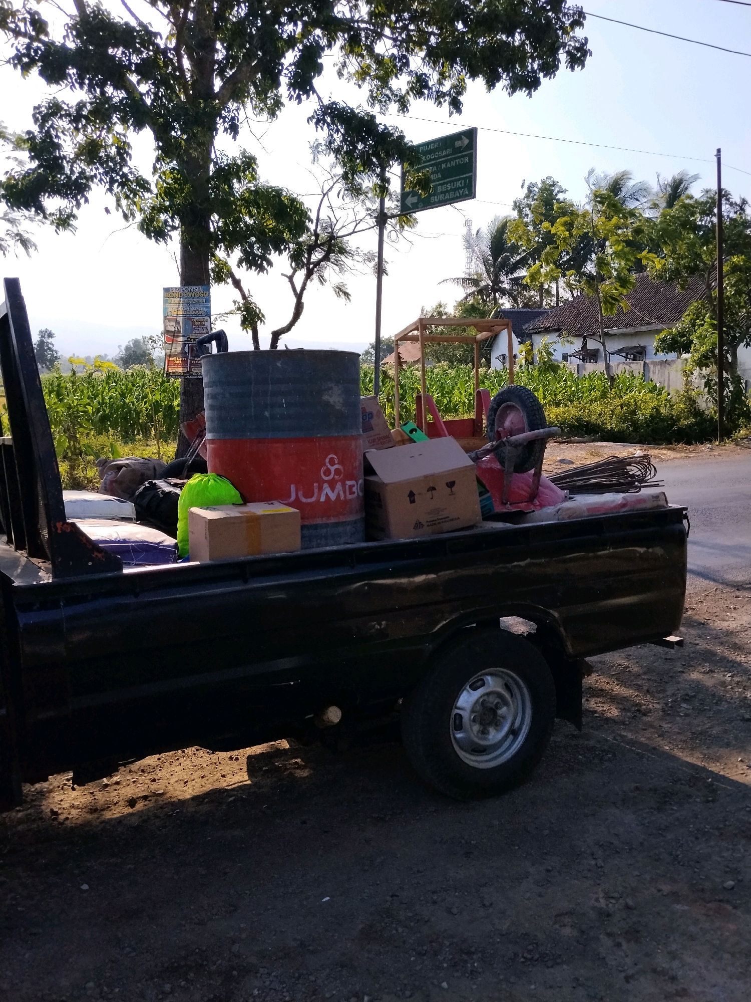A black pickup truck is parked on a dirt road next to a paved road. The truck bed is filled with various items, including a large red and gray barrel with the word "JUMBO" printed on it, several cardboard boxes, a bright green bag, and what appears to be construction materials like rebar and bags of cement. A spare tire is also visible in the truck bed. In the background, there are trees, some with lush green leaves, and a few buildings with tiled roofs. A directional signpost is visible on the left, indicating directions to "PUJEN / BOGOSARI", "STA. / KANTOR", and "BESUKI / SURABAYA". The sky is bright and clear. The scene appears to be set in a rural or semi-rural area, possibly in or near Kejawan, Indonesia, given the directional signage. The lighting suggests it is daytime, possibly late afternoon due to the long shadows. There are no people visible in the image.