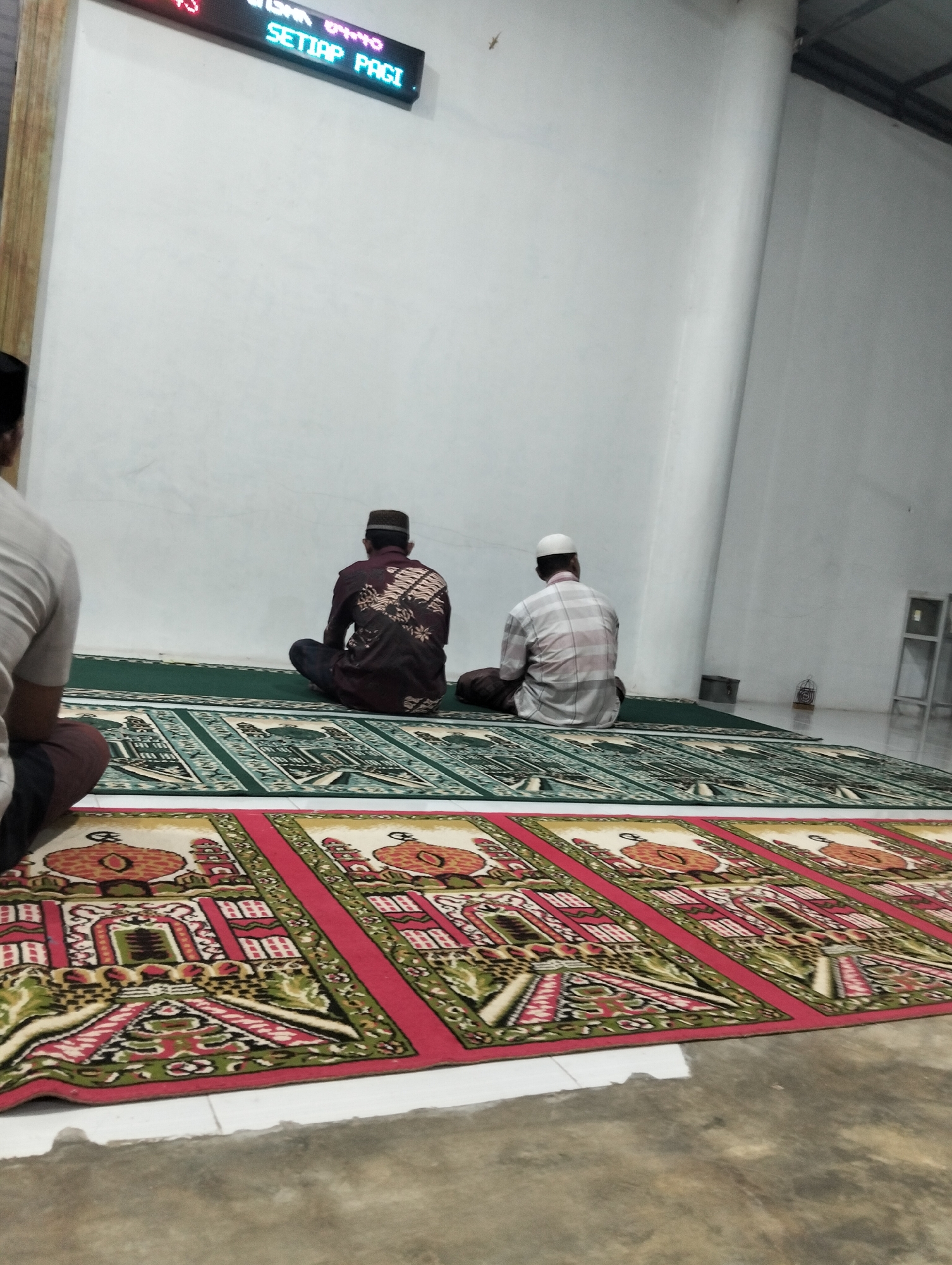 In Payagaboh, Indonesia, the image captures an interior scene of what appears to be a mosque or prayer hall. Two men are seated on prayer mats, facing away from the camera. The man on the left wears a dark shirt with a patterned design and a kufi cap. The man on the right is wearing a checkered shirt and a white skullcap. Both are in a seated prayer posture. The floor is covered with prayer rugs, some featuring geometric patterns and depictions of mosque-like structures in red, green, and yellow. A digital display board is mounted on the wall above, showing text in blue. The walls are painted white, and the lighting suggests an indoor setting, possibly during the day or evening. The overall environment is clean and orderly, indicative of a place of worship.