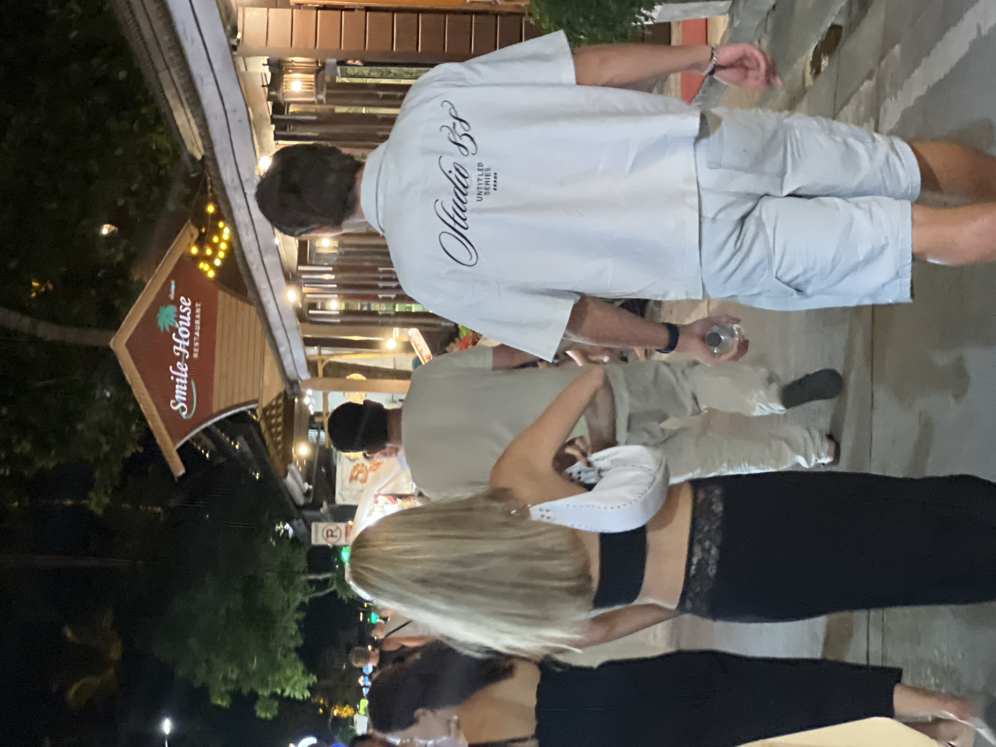 The image depicts a nighttime scene in Baan Bo Phut, Thailand, likely a street or walkway outside a restaurant. The lighting suggests it's evening or after dark, with strings of warm-toned lights illuminating the area.
Several people are visible, mostly seen from the back or side. In the foreground, a woman with long blonde hair is wearing a black top with lace detailing and black pants. She is holding a white purse. To her left, another woman's back is visible.
Further back, a man in a white short-sleeved shirt and light-colored shorts is walking away from the camera. His shirt has a graphic design that reads "Studio SS" with "UNTITLED SERIES" below it. He is holding a clear glass or bottle. Another person in a light-colored top is visible in the background.
A prominent sign for "Smile House RESTAURANT" is mounted on a wooden structure to the left, with a stylized palm tree graphic. The overall atmosphere appears casual and social, consistent with a restaurant or entertainment district. The weather seems to be clear, allowing for outdoor activity.