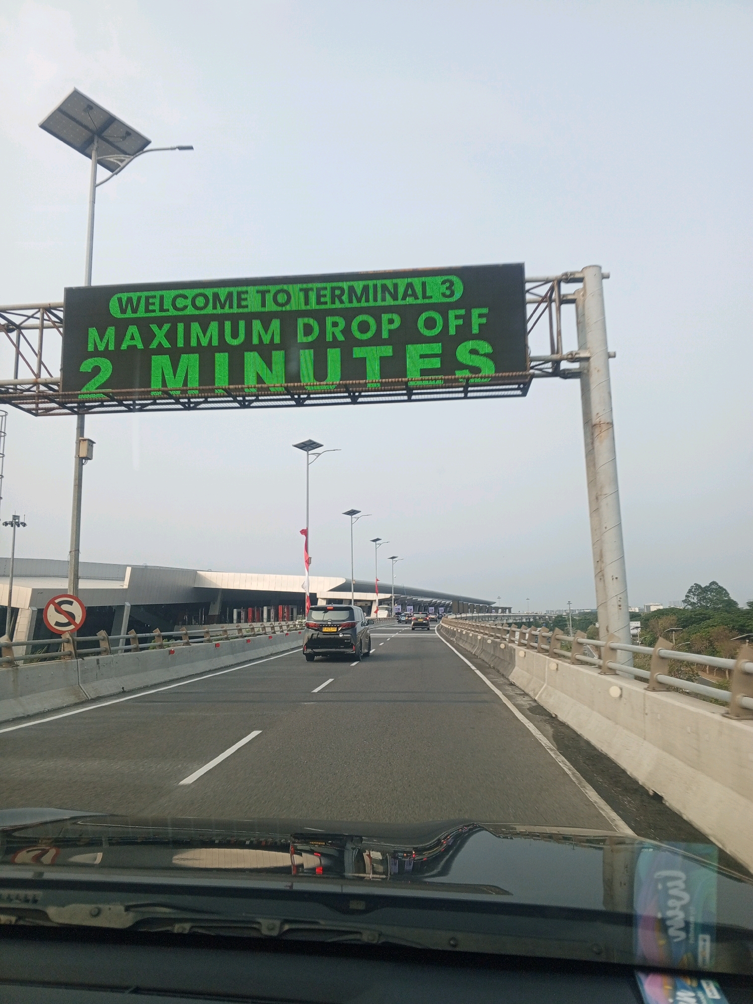 The image shows a view from a car on a highway approaching an airport terminal. A large electronic sign above the highway displays "WELCOME TO TERMINAL 3" and "MAXIMUM DROP OFF 2 MINUTES" in green LED lights. Several cars are on the highway, and a black SUV is in the right lane. The airport terminal building is visible in the background. The scene is set in Tangerang, Indonesia, likely near Soekarno-Hatta International Airport. The sky is overcast.