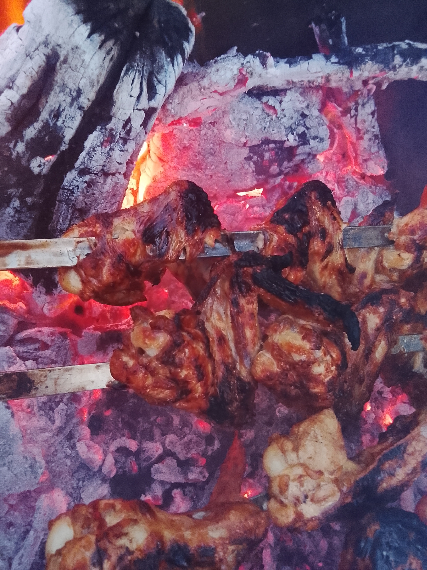 In Bondarevskii, Russia, a close-up, overhead shot captures chicken wings being grilled over hot coals. The image shows metal skewers holding the chicken as it cooks, with flames and embers visible beneath. The chicken appears to be well-seasoned and slightly charred in some areas. The background consists of burning logs and ash, indicating an outdoor cooking environment.
