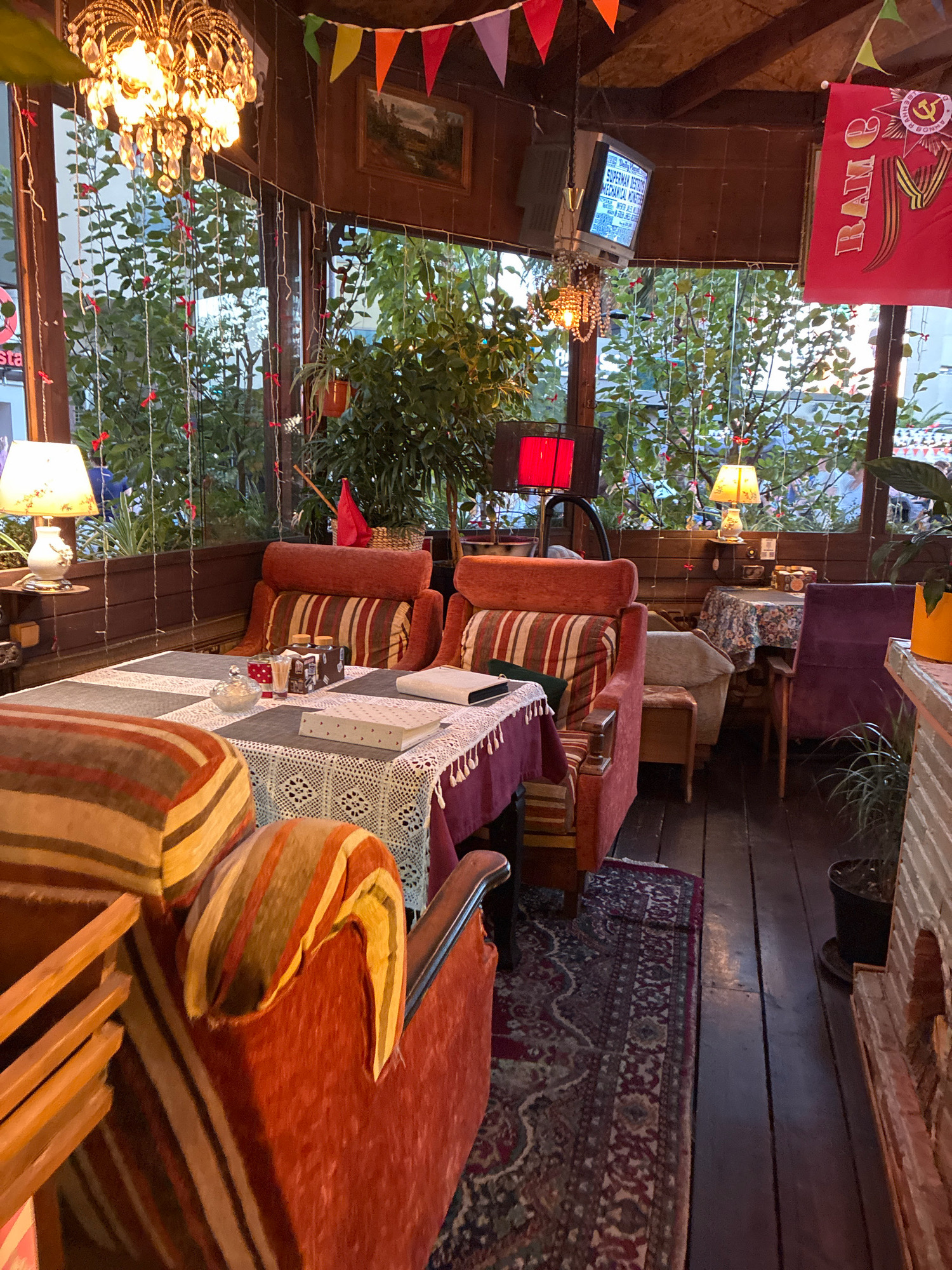 This is an interior shot of a cozy, retro-themed cafe, likely located in Sochi, Russia. The decor suggests a vintage aesthetic with plush, striped armchairs and sofas arranged around tables covered in lace tablecloths. A ornate crystal chandelier hangs from the ceiling, casting a warm glow. String lights and colorful triangular flags add to the festive atmosphere. Large windows offer a glimpse of greenery outside, with plants visible both indoors and through the glass. A small television screen displays text, and a red flag with a Soviet emblem is prominently displayed on the wall. The overall impression is one of comfort and nostalgia, creating an inviting space for patrons.