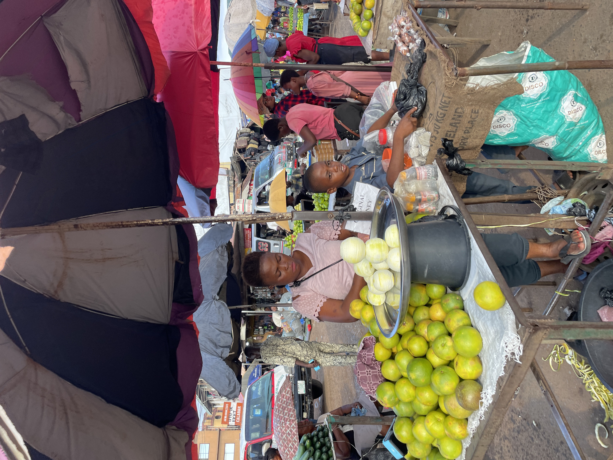 In Nkpor, Nigeria, an outdoor market scene unfolds under colorful umbrellas. A woman, wearing a pink top, stands behind a display of oranges. In front of her, a young boy in a blue shirt sits on a stool, his hands near a variety of items on a table. Behind them, other vendors and customers are visible, with vehicles and stalls suggesting a bustling marketplace. The ground is dirt, and the overall atmosphere is that of a lively street market.