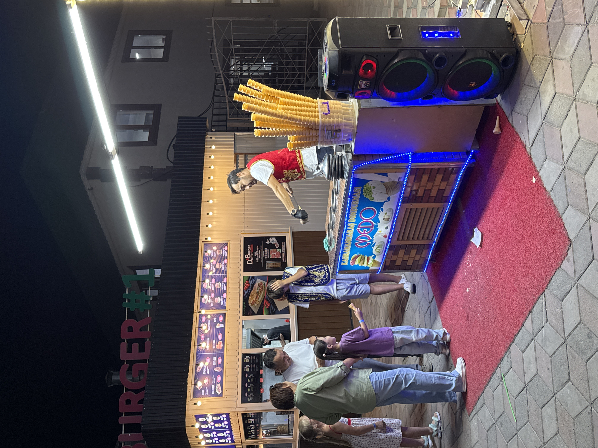 The image depicts an outdoor scene at night, likely in an urban setting. A burger joint, identified by a prominent sign reading "BURGER #1", is the central focus. The establishment features a counter with a menu displayed above, showcasing various food items. Several people are gathered around, appearing to be customers. A man in a red vest is actively serving customers, presumably at an ice cream cart which is decorated with bright blue lights and displays images of ice cream cones. A tall speaker with colorful lights is placed next to the ice cream cart. The ground is paved with interlocking hexagonal tiles, and a red rug is laid out in front of the ice cream cart. The lighting suggests it's evening or night, with artificial lights illuminating the scene. The overall atmosphere is that of a casual food stall or eatery operating in a public area.