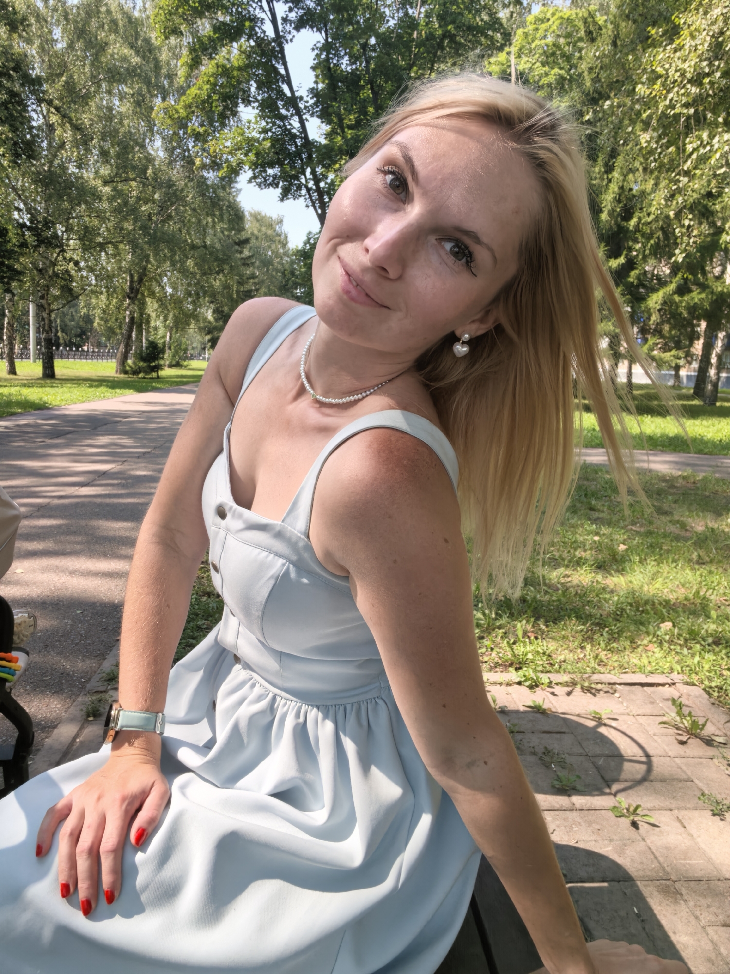 A blonde woman with green eyes and bright red nail polish is seated on a park bench, outdoors. She is wearing a light blue dress and a pearl necklace. Her head is tilted back slightly, and she is looking at the camera with a slight smile. The background of the image consists of trees, grass, and a paved path, suggesting a park setting. The lighting indicates it is daytime, with sunlight filtering through the trees. The location is identified as Sterlitamak, Russia.