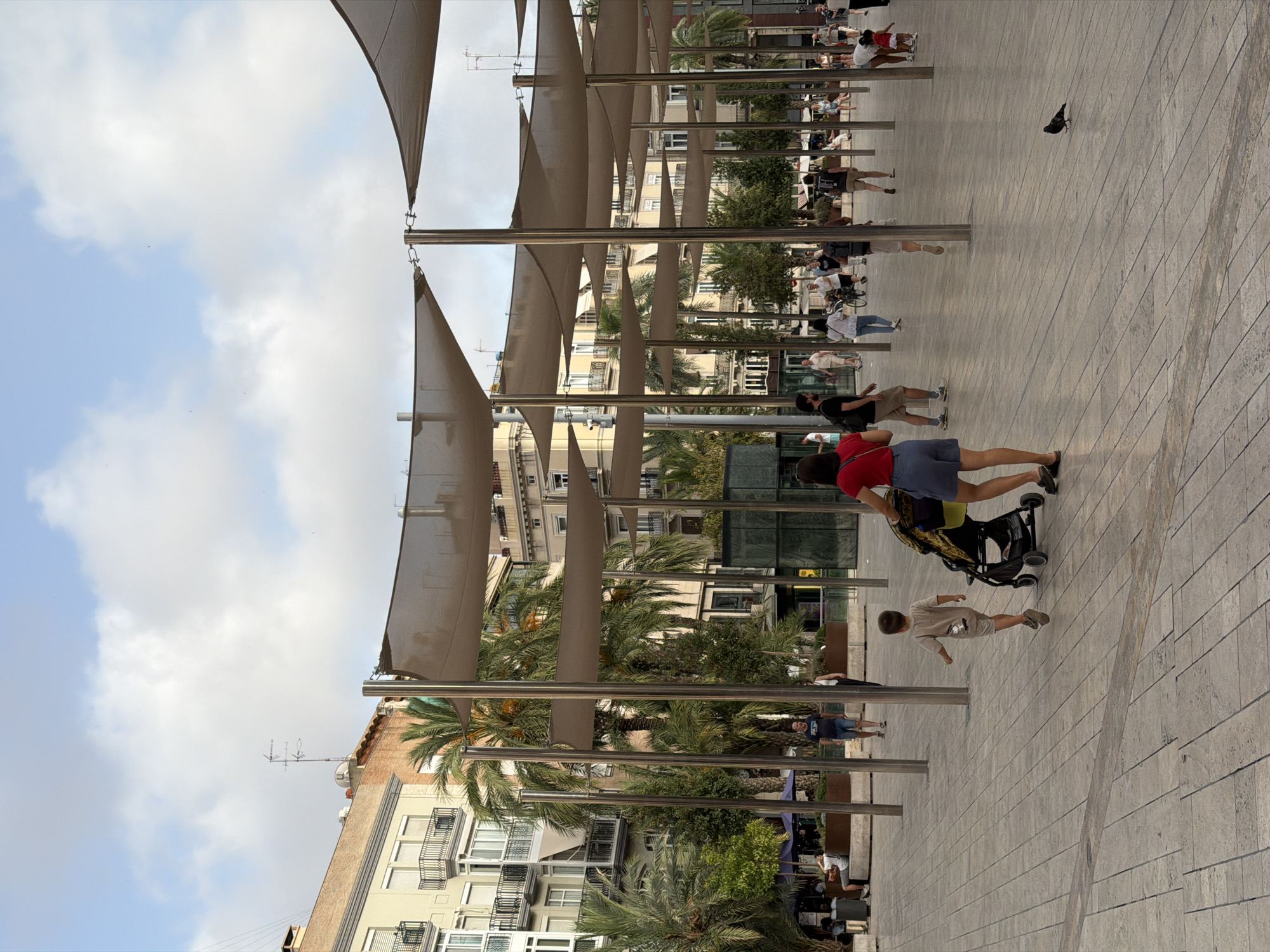 The image depicts an outdoor public space in a city, characterized by a paved ground surface and a canopy of fabric shades supported by metal poles. Several palm trees are scattered throughout the scene, adding to the tropical or Mediterranean feel. Buildings with multiple stories, featuring balconies and windows, line the background, suggesting an urban environment.
In the foreground, a young child in a light-colored outfit is walking away from the camera, towards a stroller being pushed by an adult woman wearing a red top and a blue skirt. The adult appears to be looking forward.
Further into the scene, a diverse group of people are walking and interacting. Some are walking in pairs or alone, while others are gathered in small groups. A man in a blue t-shirt and shorts is walking towards the left, facing away from the camera. Another man in a dark t-shirt and shorts is walking towards the right. A woman in a white top and blue jeans is pushing a bicycle. Other individuals are seen in the background, some appearing to be seated on benches or in conversation.
A pigeon is visible on the ground in the mid-ground, facing left. The sky above is partly cloudy, with patches of blue sky visible between the clouds. The overall atmosphere appears to be casual and relaxed, suggesting a typical day in a public square. The architecture and vegetation indicate a warm climate.