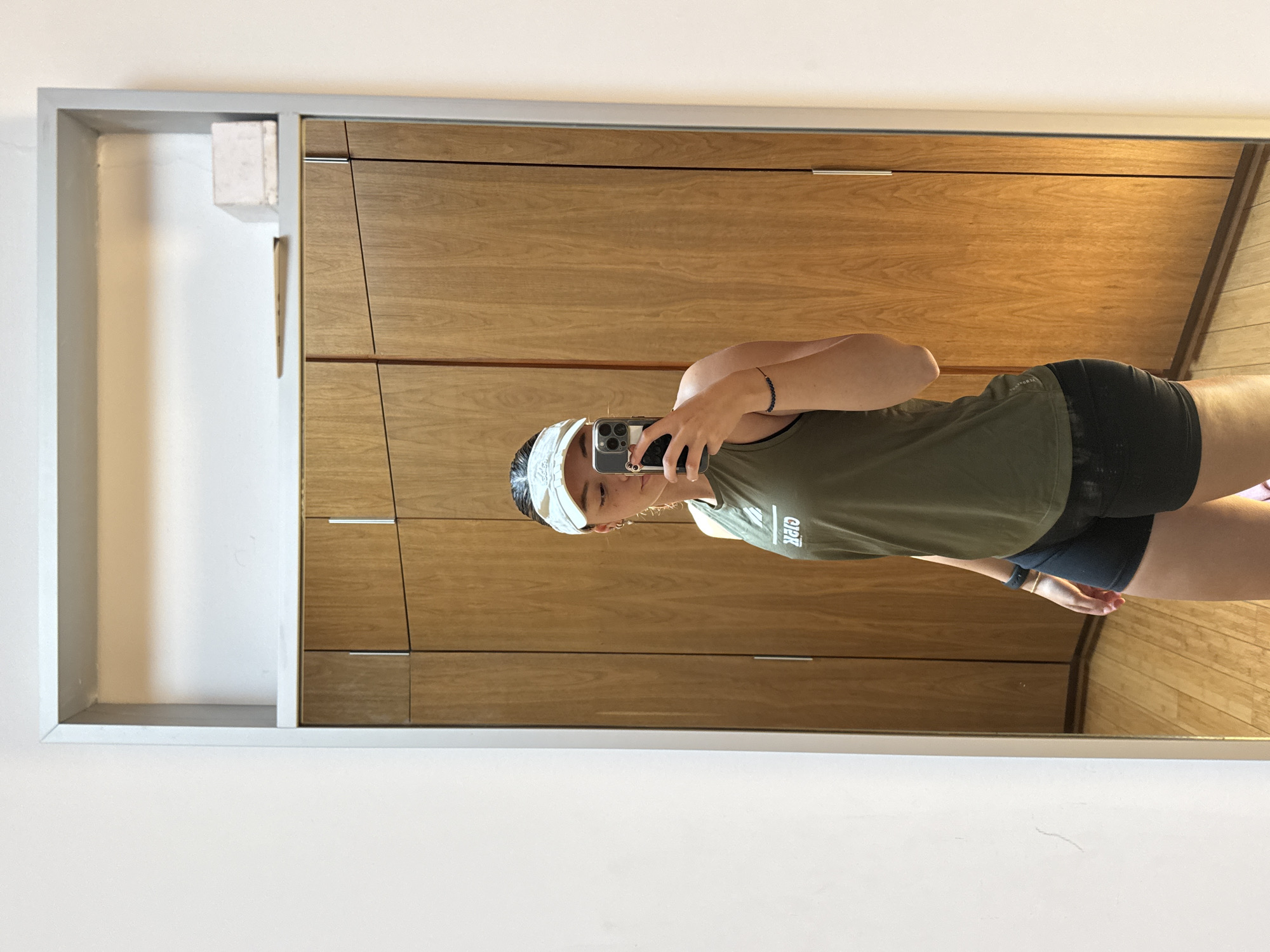 A woman is taking a selfie in front of a mirror. The mirror is framed in gray and reflects a section of the room, showing light brown wooden wardrobe doors and a glimpse of a wooden floor. The woman is wearing a white visor, an olive-green tank top, and black shorts. She is holding a smartphone up to take the photo. The location details are not discernible from the visual information.