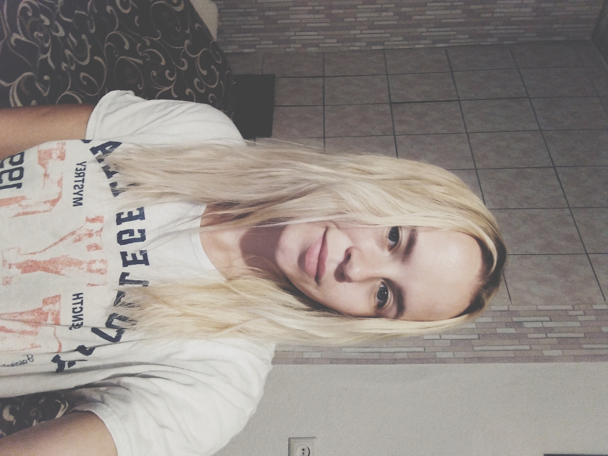 A young woman with blonde hair and light skin is taking a selfie in an indoor setting. She is wearing a white t-shirt with a graphic design on it. The background shows a tiled wall and a bed with a patterned comforter. A power outlet is visible on the wall to the right. The image appears to be taken in a home environment. Radisne, Ukraine is not identifiable from the image.