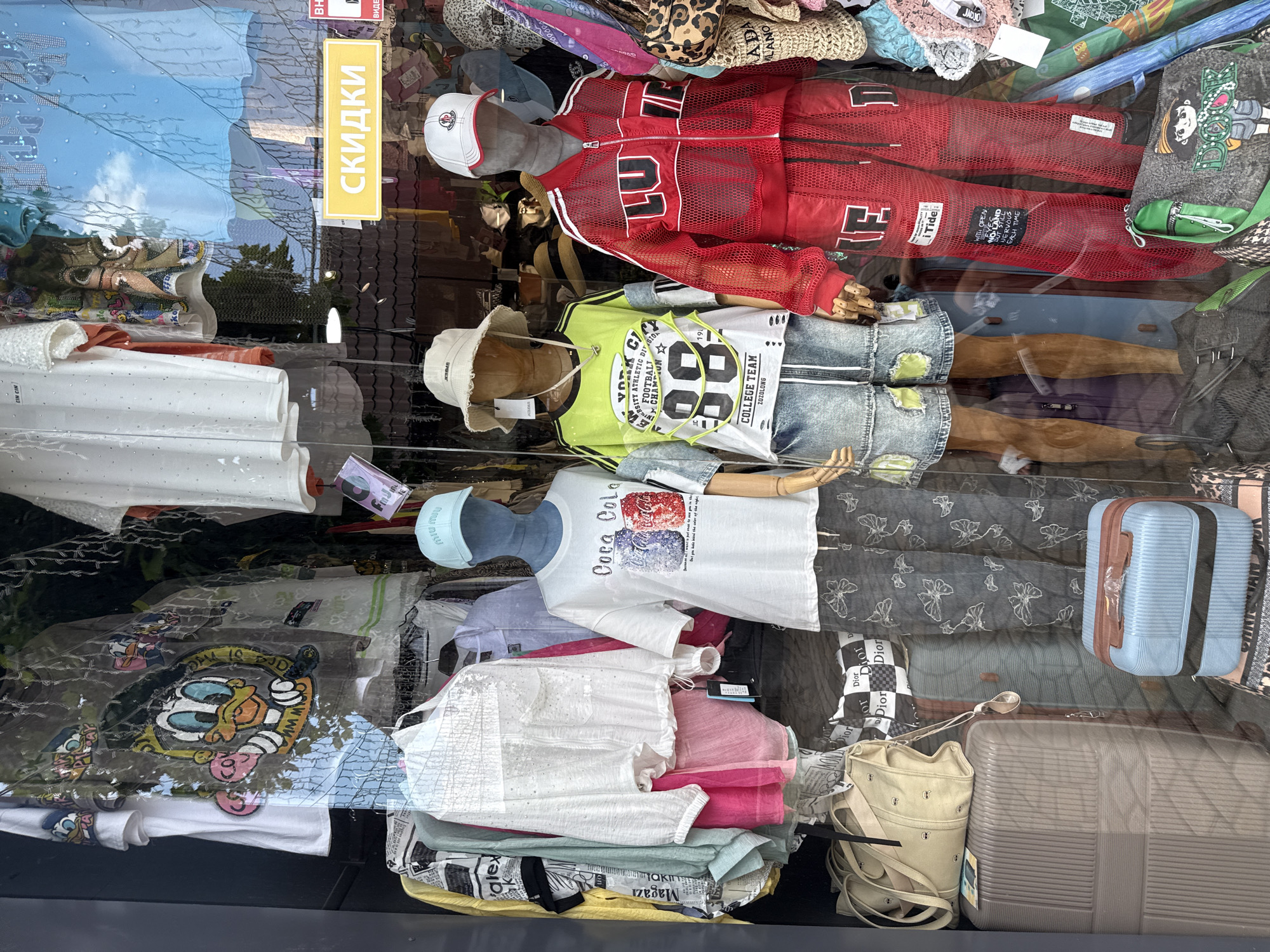 This image captures a street-level view of a retail shop window display in Sochi, Russia. The display features an array of clothing and accessories arranged on mannequins and racks. Prominently displayed are several t-shirts, including one with a "Coca-Cola" graphic and another with a cartoon character. There are also shorts, pants, a red mesh tracksuit, and a white sequined shirt. Accessories such as hats, bags, and luggage are also visible. A yellow sign with Russian text, "СКИДКИ," which translates to "Discounts," is affixed to the window. The reflection in the window suggests an outdoor environment with sky and trees visible. The overall scene depicts a commercial setting focused on fashion retail.