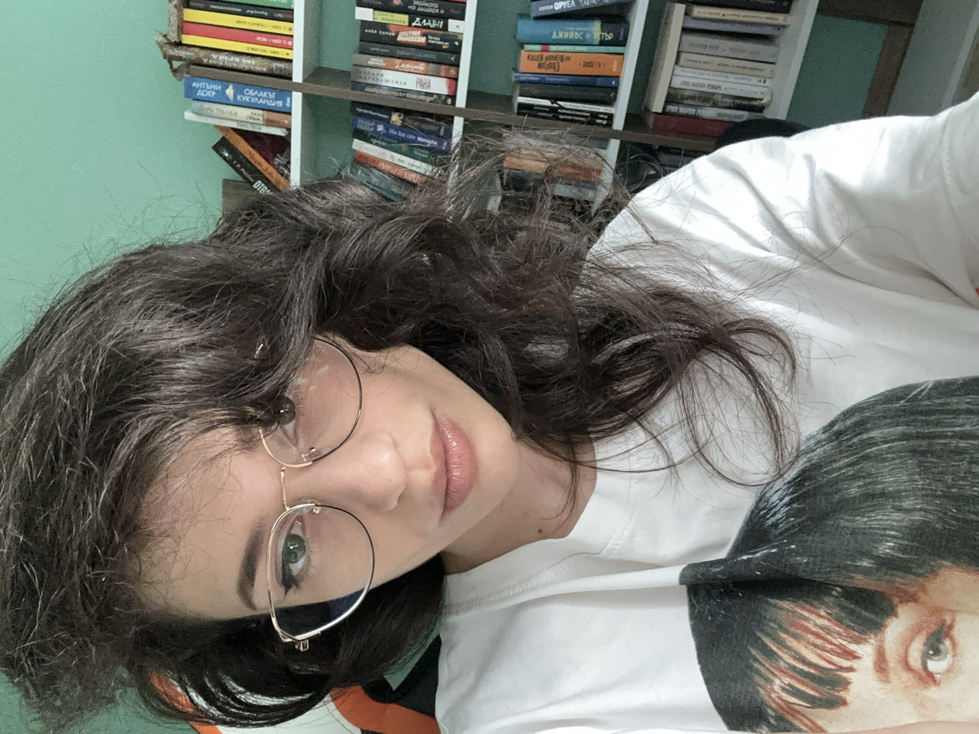 The image shows a close-up, selfie-style shot of a young woman wearing a white t-shirt with a printed image of a woman's face. She has dark, wavy hair, light green eyes, and is wearing round, gold-rimmed glasses. Her lips are slightly parted. In the background, a bookshelf filled with books is visible, and the wall behind it is painted a light green color. The image is taken indoors, likely in a residential setting, and there is no indication of the specific location within the city of Sofia, Bulgaria.
