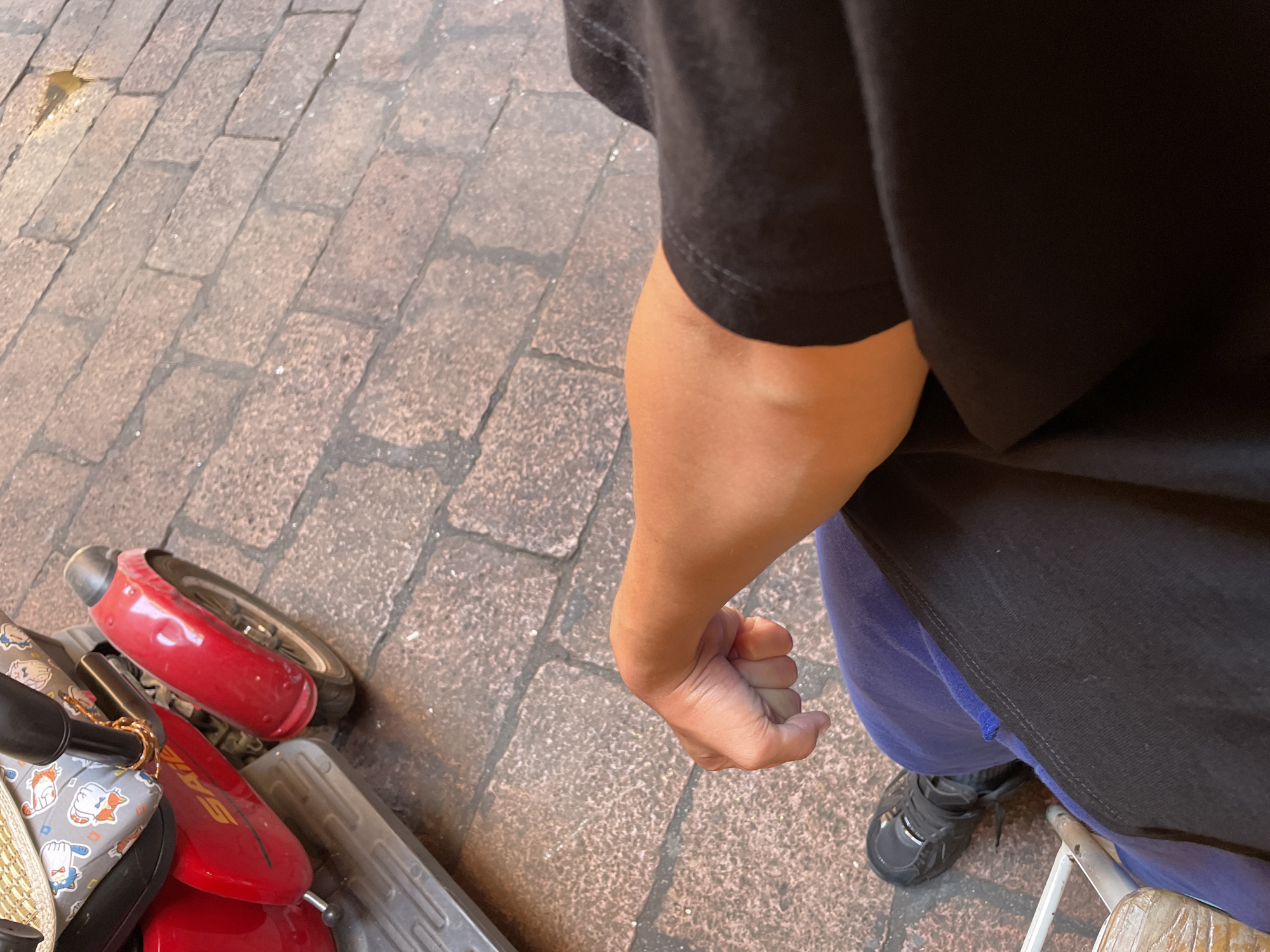 The image depicts a person standing on a brick-paved surface, presumably a sidewalk or pedestrian area in the city of Bologna, Italy. The person is wearing a black t-shirt and blue pants. A black shoe is visible. A small scooter or mobility device is in the lower left of the frame. The person's hand is clenched. The lighting suggests it is daytime.