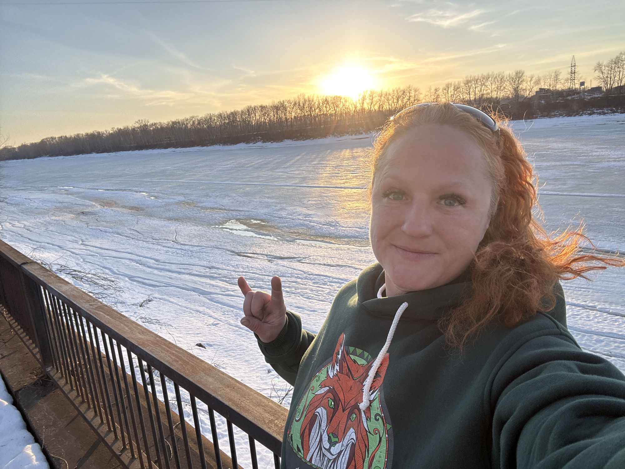 The image is a selfie of a woman with red hair wearing a green hoodie with a fox design. She is standing next to a metal railing. In the background is a wide, frozen river with a line of trees on the opposite bank. The sun is setting behind the trees, casting a bright light on the scene. Based on the presence of the frozen river, the location is likely Chaikovsky, Russia.