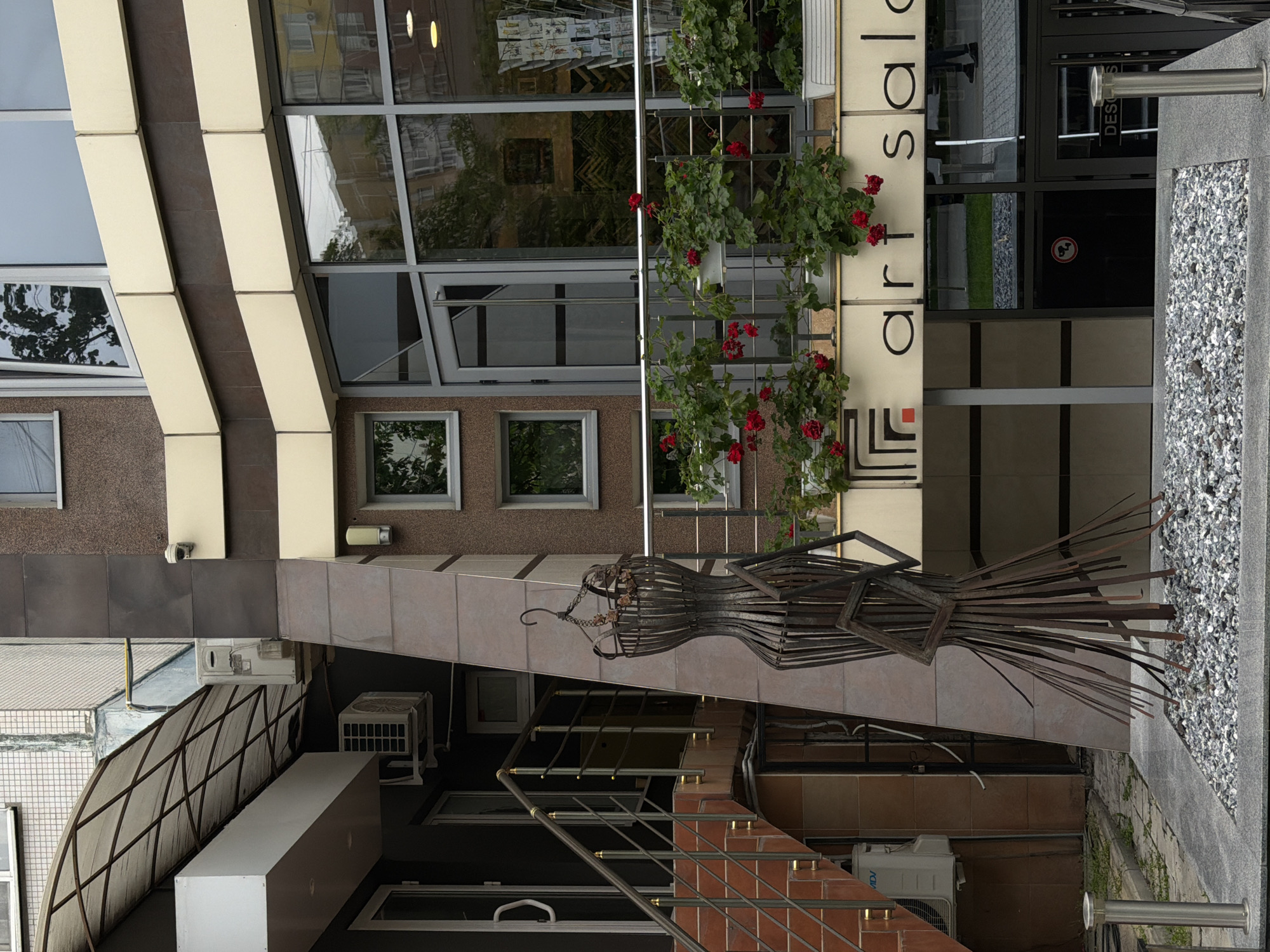 The image shows the exterior of a building, likely a commercial space. A decorative metal sculpture of a dress form is mounted on the building's facade. Above the sculpture, the words "art salon" are visible on a sign, flanked by red flowering plants. The building features large windows and a modern architectural design with alternating beige and brown panels. Below the sculpture, there is a staircase with a metal railing, and an air conditioning unit is visible. The ground is covered in gravel. The location is in an urban area, although the city is unknown.