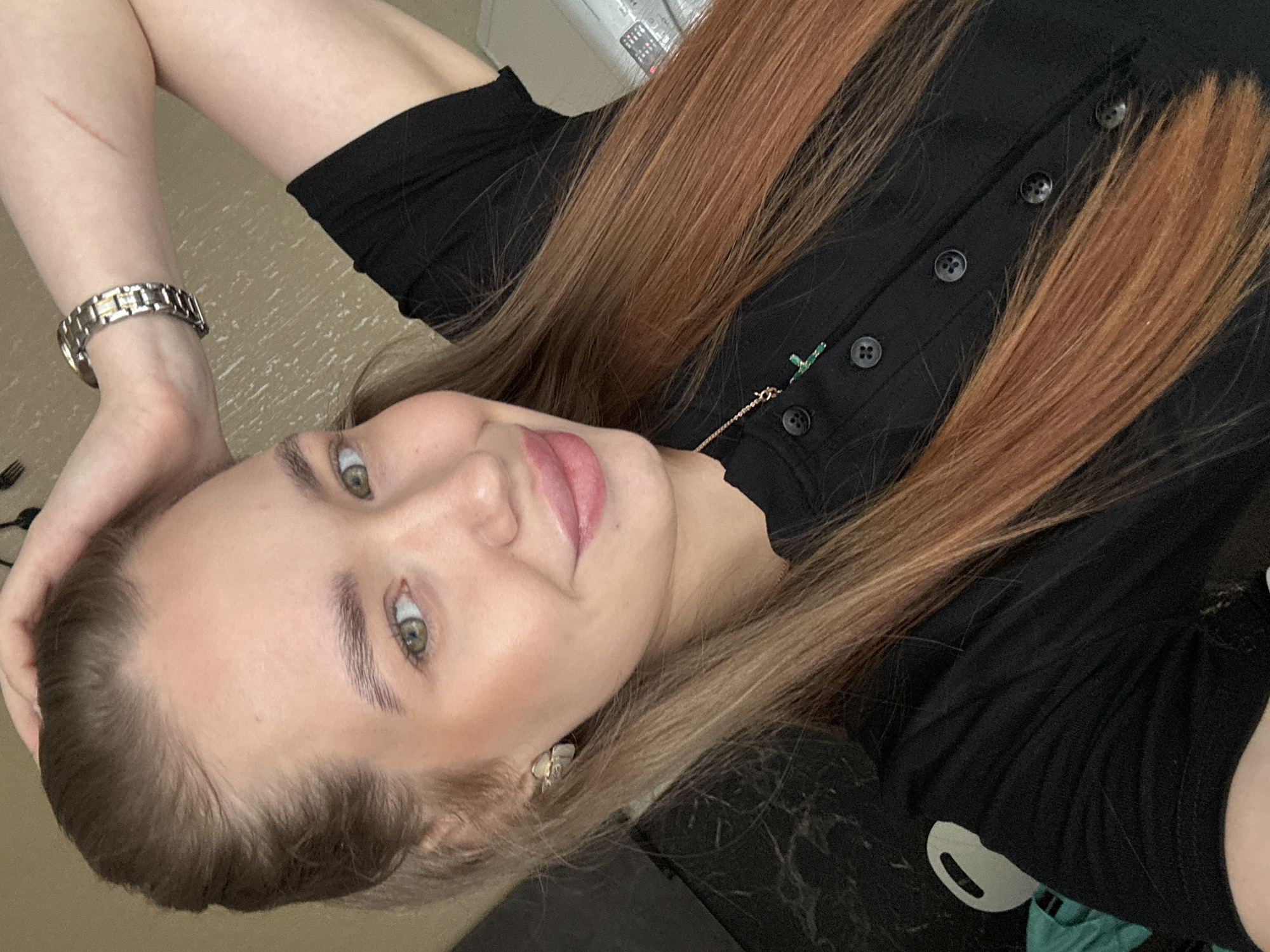 A woman with long reddish-brown hair is pictured. She is wearing a black top with buttons and a delicate necklace. A silver watch adorns her left wrist. Her face is slightly turned, revealing her eyes and a gentle smile. An earring is visible in her left ear. Her left hand is near her head. The background is blurred and indistinct, making it difficult to determine her exact location in Chaikovsky, Russia.