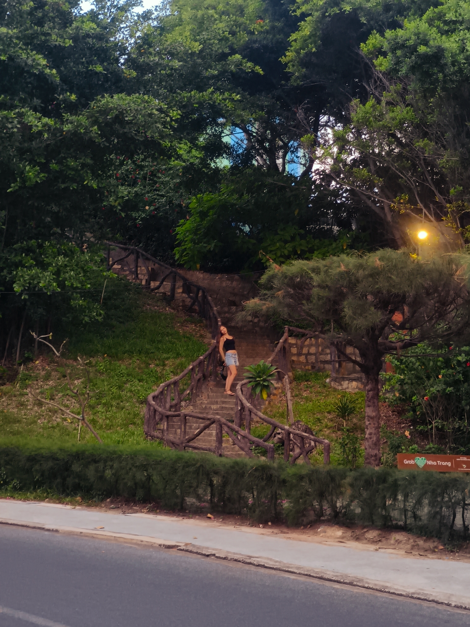 A woman is standing on a concrete staircase with a rustic wooden railing. She is wearing a black tank top and denim shorts. The staircase is surrounded by lush greenery, including trees and grass. In the background, there are more trees and some hints of blue sky. A street is visible in the foreground with a hedge separating it from the park area. A street lamp is also visible, casting a warm glow. A sign that reads "Grab Nha Trang" is present on the right side of the image.