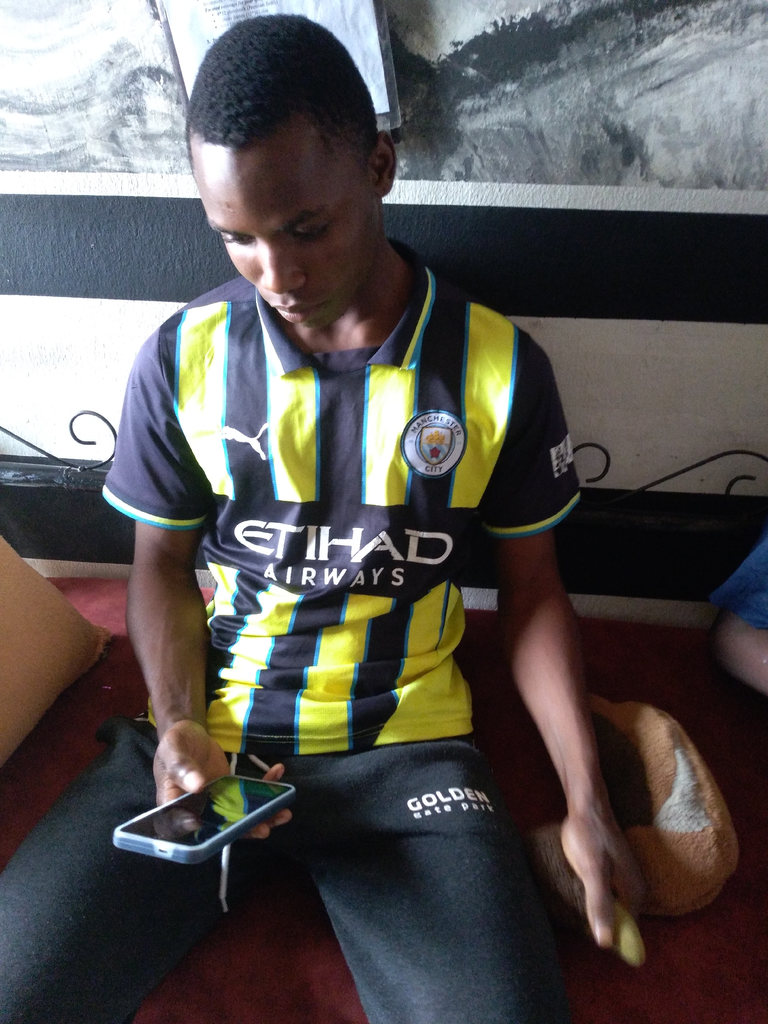 The image shows a young man sitting indoors, holding a smartphone. He is wearing a dark blue and yellow striped soccer jersey with "ETIHAD AIRWAYS" printed on the front, and black sweatpants with the word "GOLDEN" visible. The man is looking down at his phone. In the background, there is a black and white striped wall and some artwork. To his right, there is a person partially visible wearing a blue shirt. A brown and white cushion is on the seat next to him. The scene appears to be a casual, indoor setting.