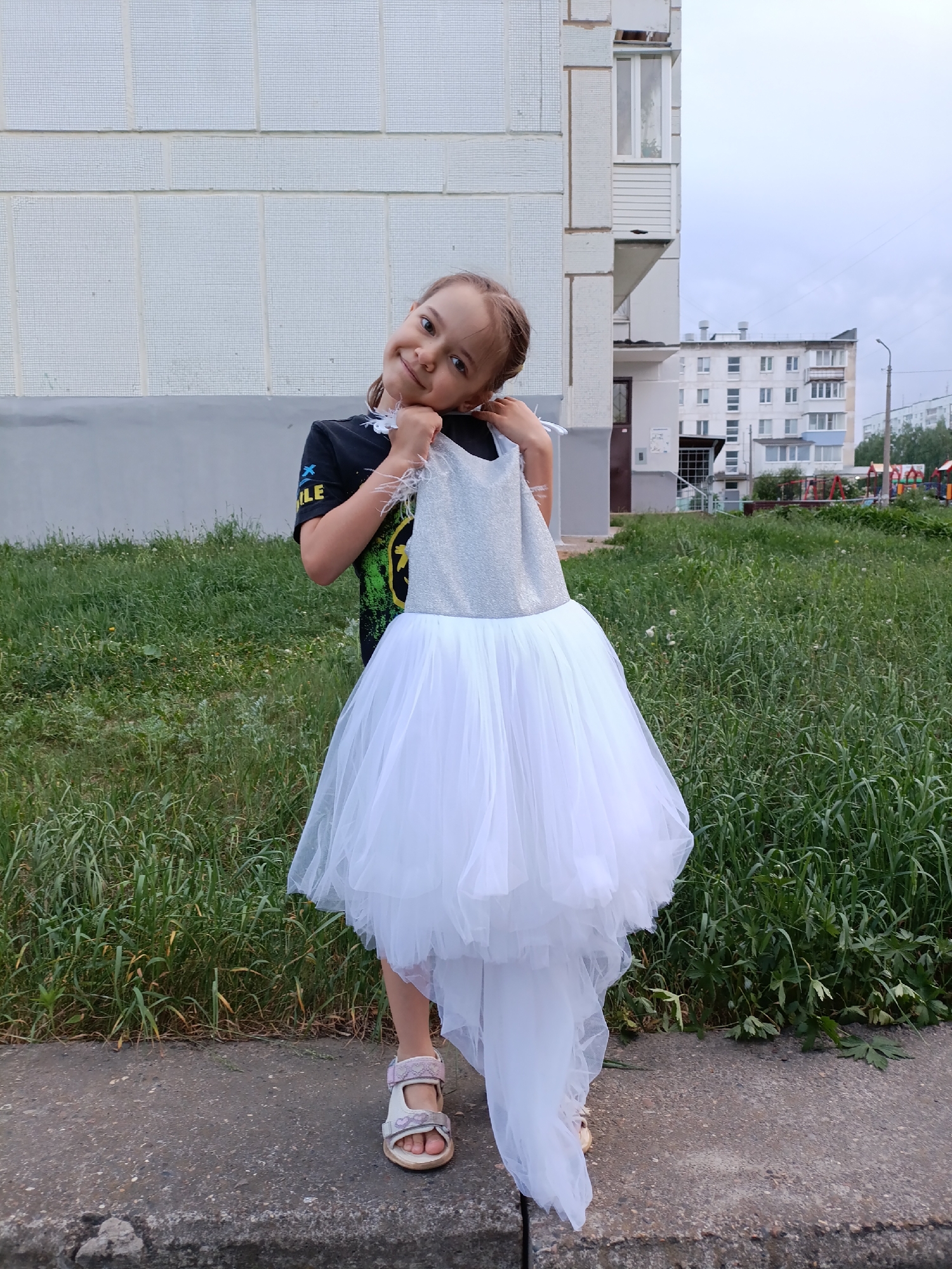 A young girl stands in front of a grassy area with buildings in the background. The girl is fair-skinned with light brown hair styled in braids. She wears a black t-shirt with a graphic and light-colored sandals. She holds a white and silver dress in front of her. The dress has a sparkly silver bodice and a layered tulle skirt with a long train. The backdrop features buildings constructed of white panels, indicative of residential architecture, possibly apartments. Further in the distance, there is a glimpse of what appears to be a playground with colorful structures. Based on the surrounding cityscape, this photo was taken in Chaikovsky, Russia.