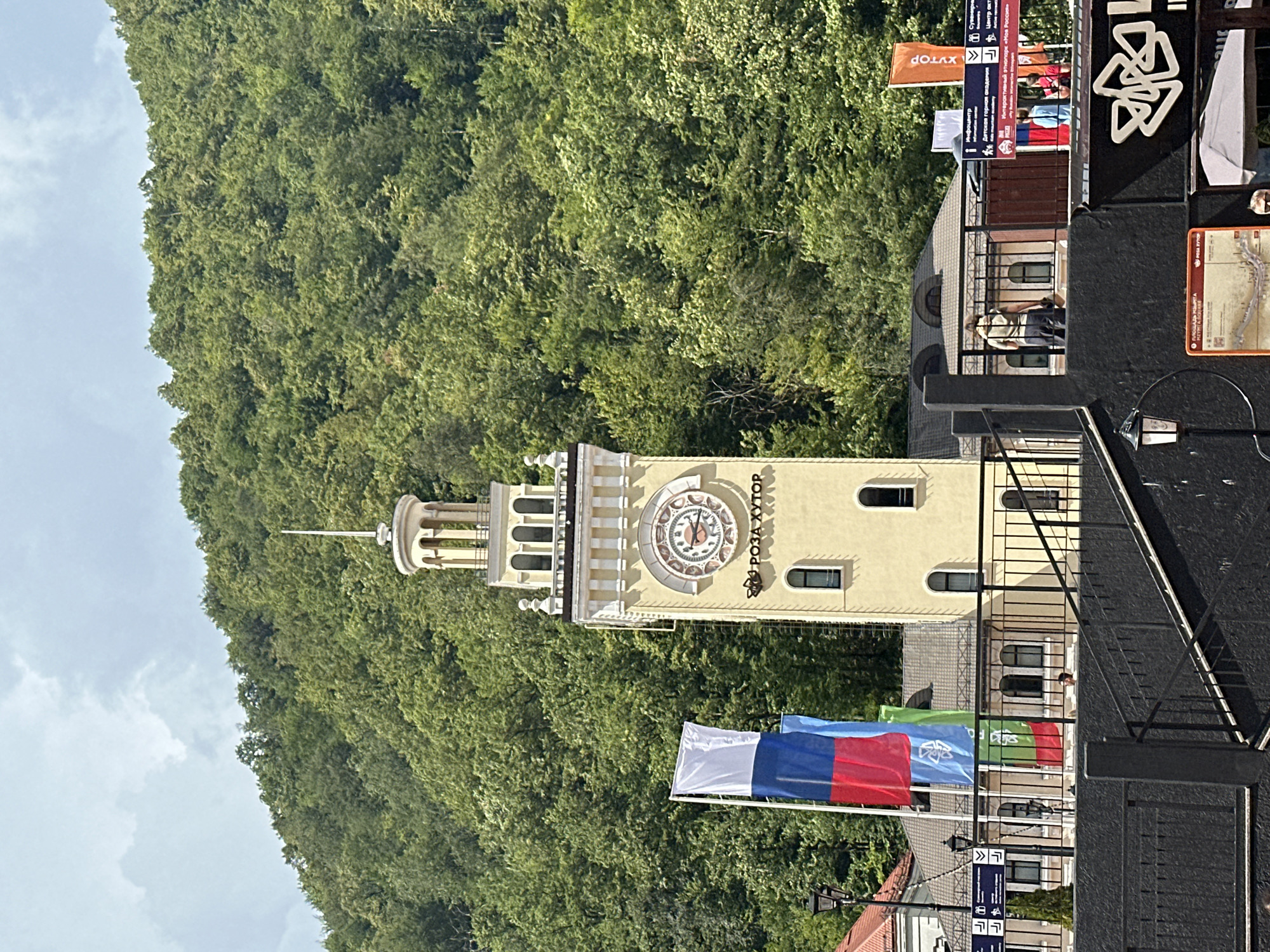 The image captures a view of the central tower and surrounding buildings in the resort town of Estosadok, Russia, nestled against a backdrop of a densely forested, green mountain slope. The tower, a prominent yellow structure with white architectural accents, features a large clock face with Roman numerals and ornate clock hands. Above the clock, the tower is crowned with a decorative cupola.
In the foreground, a black metal railing and staircase dominate the lower part of the frame, with a black wall below. To the left of the tower, the Russian flag flies prominently alongside two other flags, one featuring a blue emblem on a white background and another with a green emblem.
Behind the tower, several buildings with pitched roofs are visible. These structures appear to be commercial establishments or administrative buildings within the resort. Signs with text in Russian and pictograms are affixed to the buildings and railings, indicating directions or services.
Several individuals are present in the scene. One person is walking along a pathway behind the railing, and a few other figures are visible in the distance near the buildings. The environment suggests a clear day with scattered clouds in the sky. The overall impression is one of a well-developed tourist destination with a blend of modern infrastructure and natural beauty.