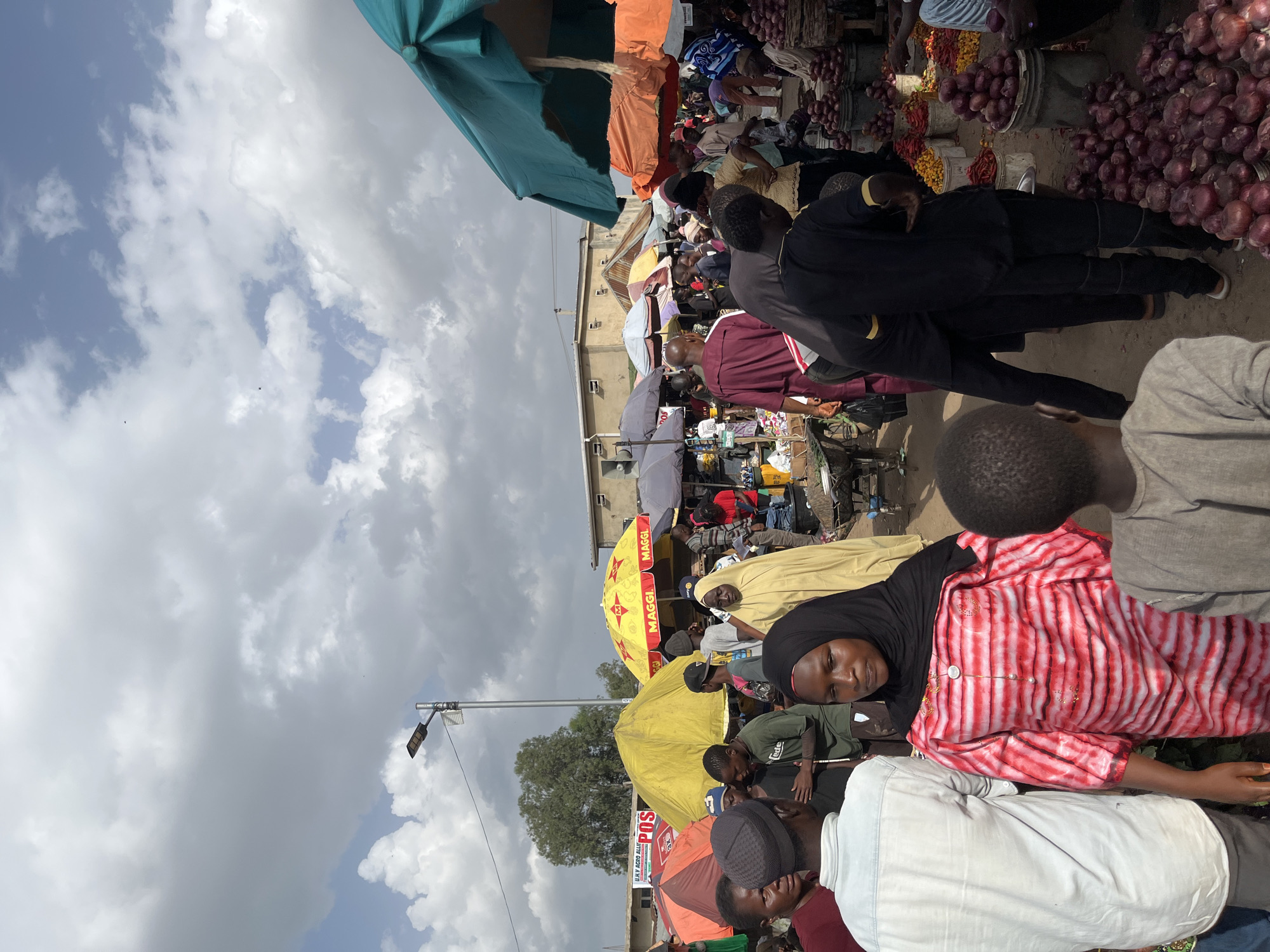 The image depicts a bustling outdoor market scene in Rafin Pa, Nigeria. A crowd of people are gathered, with many individuals browsing or interacting. In the foreground, a woman in a black hijab and a vibrantly striped red and white dress is looking to the side. To her left, a man in a light blue long-sleeved shirt and grey head covering faces away from the camera. Further back, people are seen wearing various attire, including a woman in a light yellow robe and head covering, and men in traditional Nigerian clothing like a burgundy robe. Several market stalls are visible, covered by colorful umbrellas in shades of yellow, orange, and turquoise, providing shade for vendors and shoppers. Goods for sale, primarily produce such as onions and peppers, are displayed in buckets and piles. In the background, buildings and trees suggest an urban or semi-urban environment. A street lamp is mounted on a pole above the market, indicating it is likely a public thoroughfare. The sky is partly cloudy, with a mix of sunlight and diffused light.
