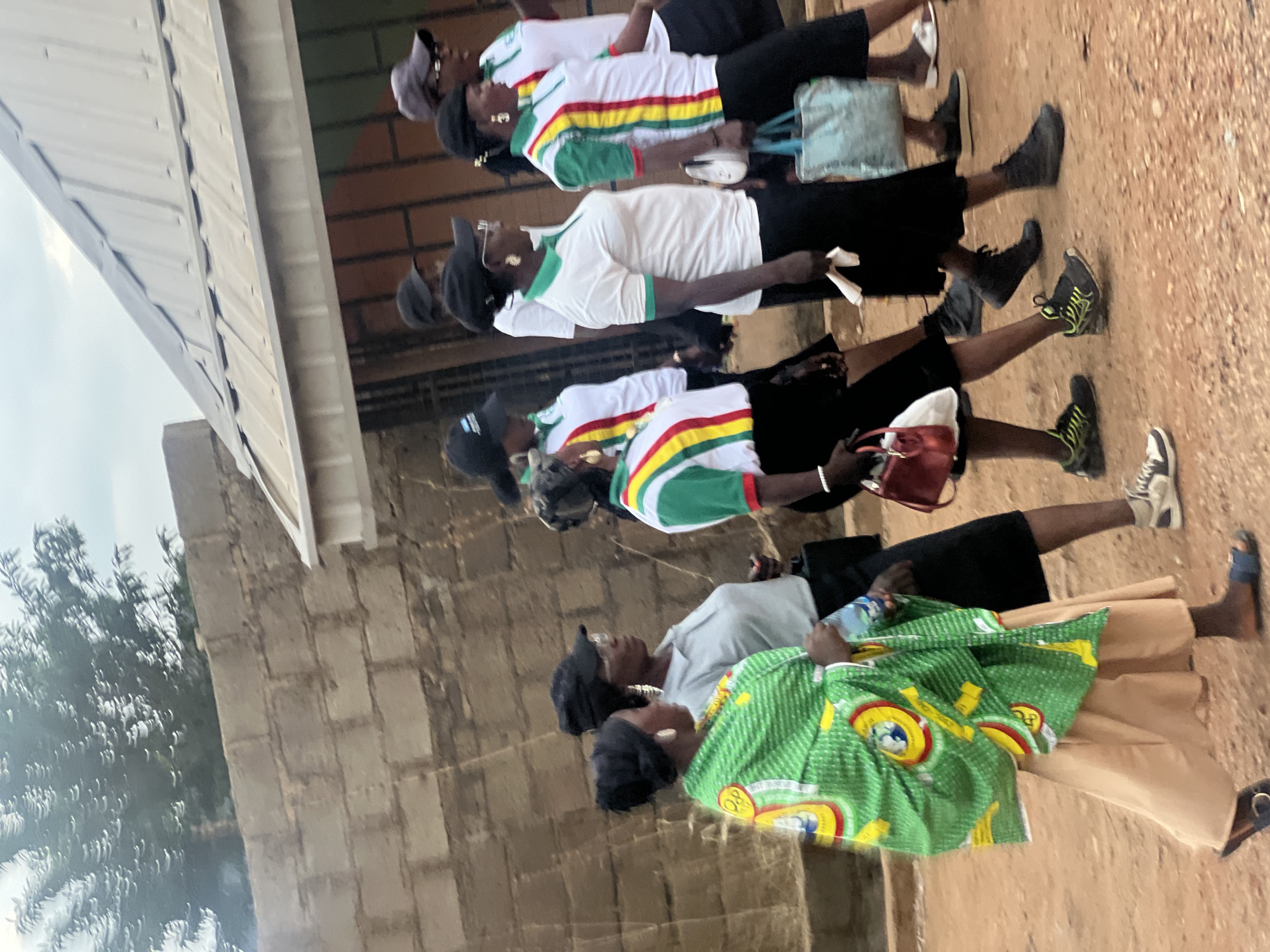The scene depicts a group of women standing outside, seemingly in Gura Top, Nigeria. Several of the women are wearing white shirts with green, yellow, and red striped accents, black skirts, and caps. One woman in the foreground wears a green patterned cloth. The ground is a dirt surface, and a building with a corrugated metal roof is visible in the background.