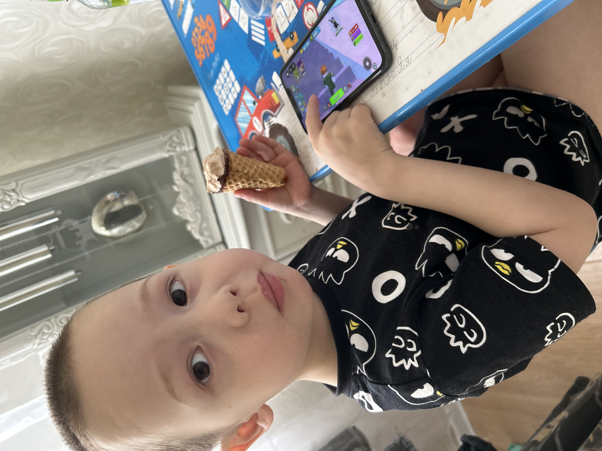 A child sits at a table, holding an ice cream cone in one hand and using a smartphone with the other, presumably to play a game. The table has a blue plastic surface with colorful graphics. The child is wearing a black shirt with white cartoon-like designs. The location is most likely a private residence in the city of Chaikovsky, Russia, as indicated by the prompt.