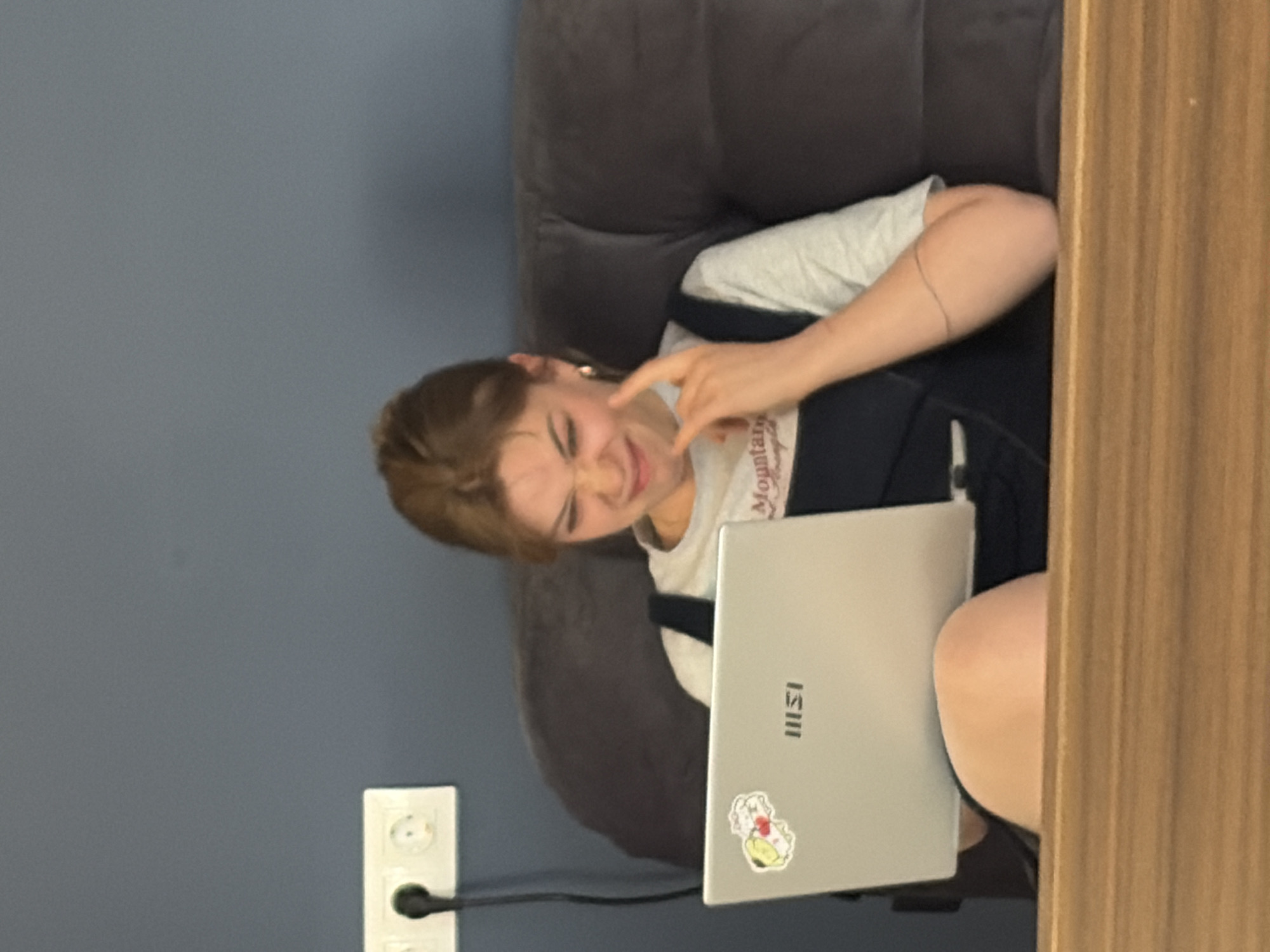 A young woman sits on a grey couch in a room in Бараўлянскі сельскі Савет, Belarus, working on a silver MSI laptop. She is wearing a grey t-shirt with the word "Mountain" visible and black overalls. She has a sticker on her laptop. Her expression suggests frustration or discomfort as she touches her face. An electrical outlet is visible on the blue wall next to the couch. A wooden panel is partially visible on the right side of the image.