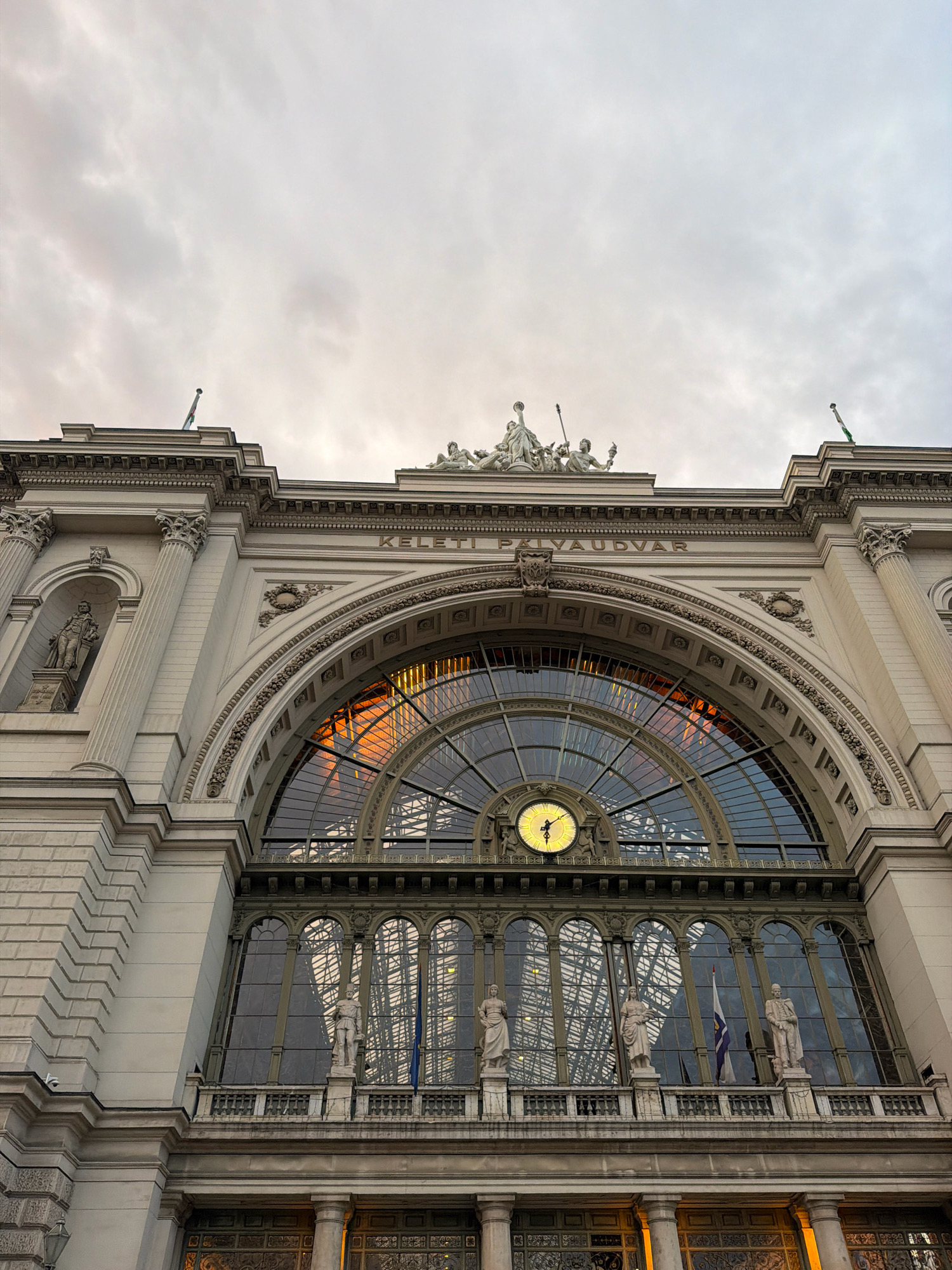 The image shows the Keleti Railway Station in Budapest, Hungary, viewed from a low angle, looking upwards at its facade. The sky above is overcast with gray clouds. The station's architecture is grand, featuring ornate stonework, columns, and statues. A large, arched glass window dominates the center of the facade, revealing the station's interior structure and reflecting the sky. A prominent clock face is visible within this arched window. Below the main arch, there are rows of smaller arched windows and a balcony with several statues. Flags are visible on the roofline. The lighting suggests it is either early morning or late afternoon.