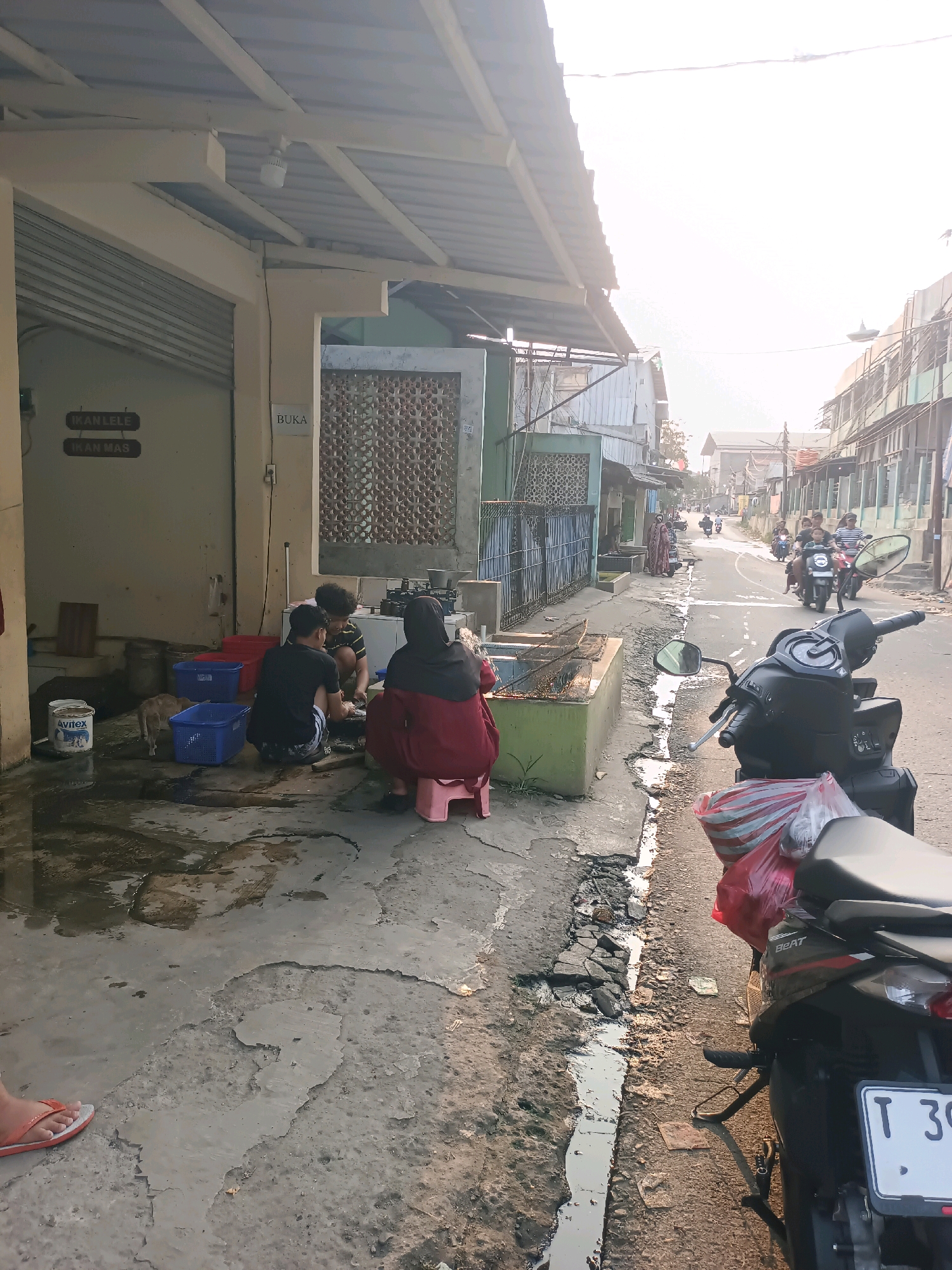 The image depicts a street scene in Karawang, Indonesia, likely near a market or food stall. In the foreground, a black motorcycle is parked on the right side of the road, partially obscuring the view of a shallow, dirty drain. To the left, several people are gathered on the ground. A woman wearing a red hijab and a maroon robe is seated on a pink stool. Beside her, two young men are squatting and appear to be cleaning fish, with a small cat nearby. Red and blue plastic baskets are placed on the wet, cracked pavement, along with a white bucket labeled "Avitex". Above them, a building with a corrugated metal roof and a sign that reads "BUKA" (Open) is visible. Further down the street, other buildings and a line of motorcycles with riders suggest a bustling urban environment. The sky is bright and overcast, indicating it might be late morning or early afternoon.