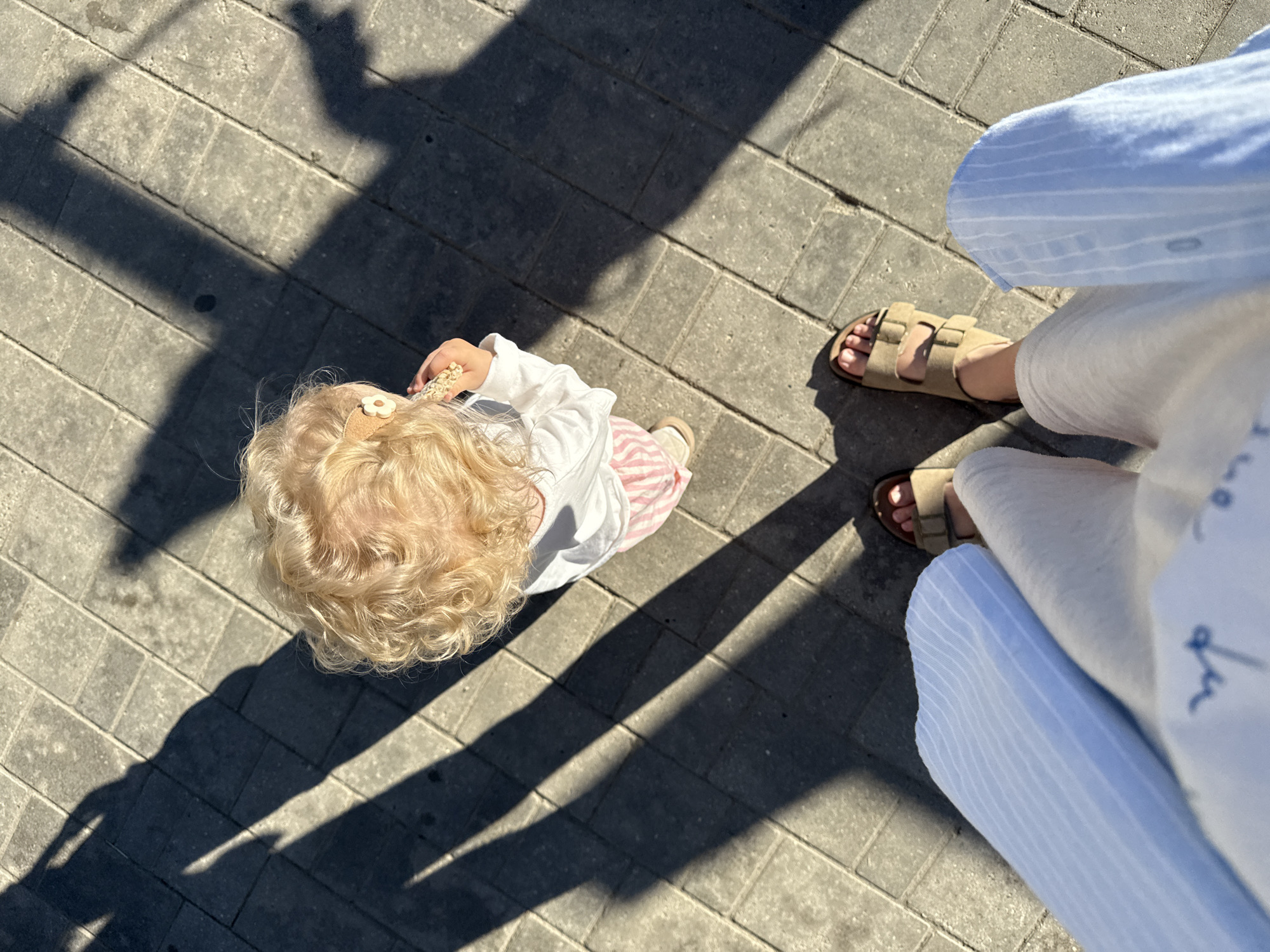 The image shows a high-angle view of a young child with blonde, curly hair walking on a paved sidewalk. The child is wearing a white long-sleeved shirt and pink and white striped pants. A small, light-colored flower clip is visible in the child's hair. The child is holding a small, light-colored object in their right hand.
To the right of the child, an adult's legs are visible, wearing light-colored pants and tan sandals. The adult is also wearing a light blue and white striped shirt. Long shadows are cast on the sidewalk, indicating that it is likely daytime and sunny. The paving stones are a uniform grey color. The scene appears to be a casual outdoor setting, possibly a park or a street in a city. There is no visible text in the image.