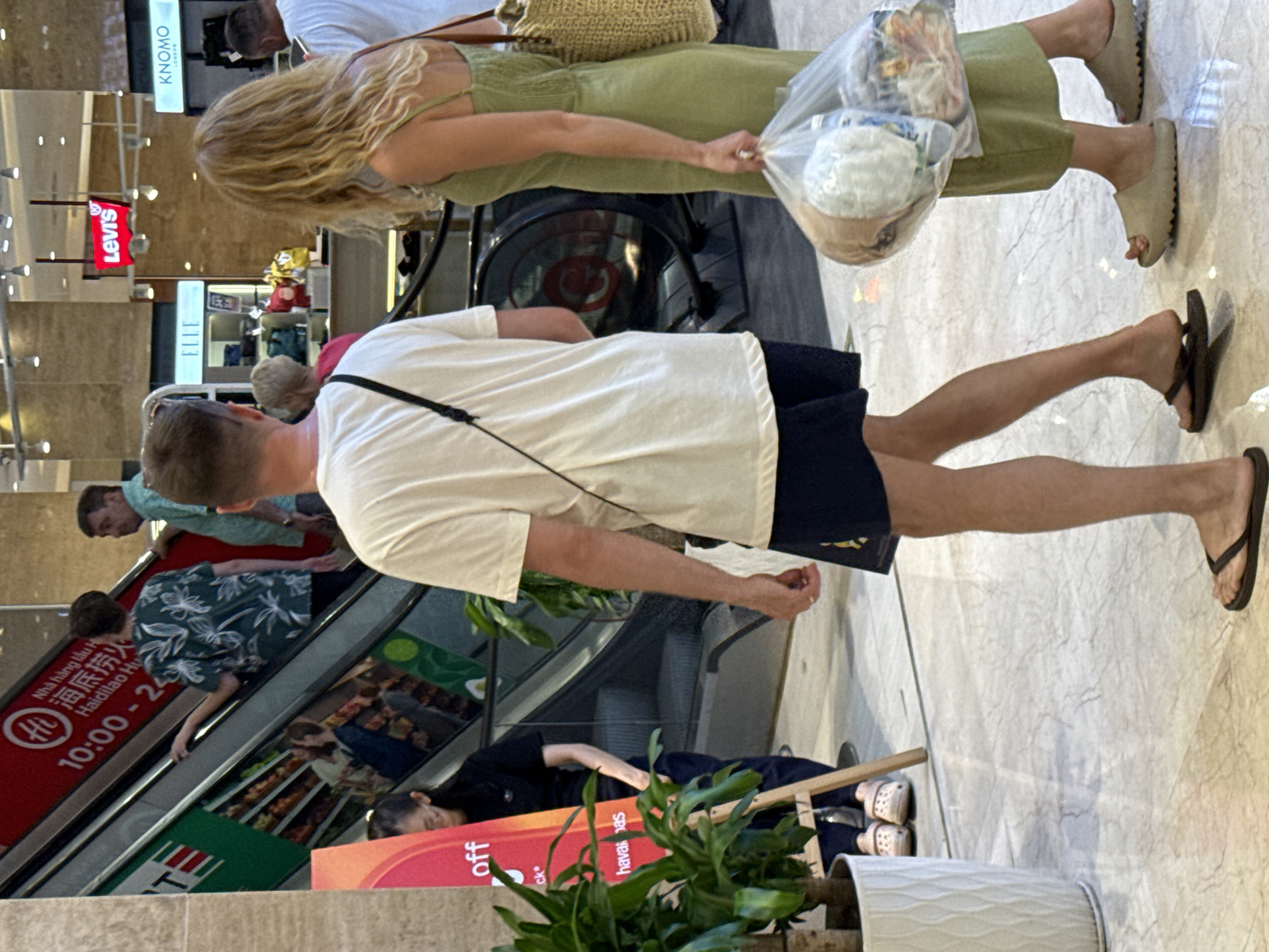 The image depicts an indoor scene, likely a shopping mall, with a prominent escalator. In the foreground, a man wearing a white t-shirt and navy shorts with flip-flops walks away from the camera, carrying a black bag slung across his chest. To his right, a woman in a light green dress walks towards the camera, holding a transparent plastic bag filled with items and wearing beige slides. Behind them, the escalator displays branding including "Hi" and Chinese characters, with the text "10:00 - 24:00" visible, suggesting operating hours. Several other individuals are visible on and around the escalator, some facing forward and others facing backward. The environment appears to be a commercial setting with polished marble flooring and store signage in the background, including "Levi's" and "KNOMO". A potted plant is situated to the left of the frame. The location is identified as Nha Trang Ward, Vietnam.