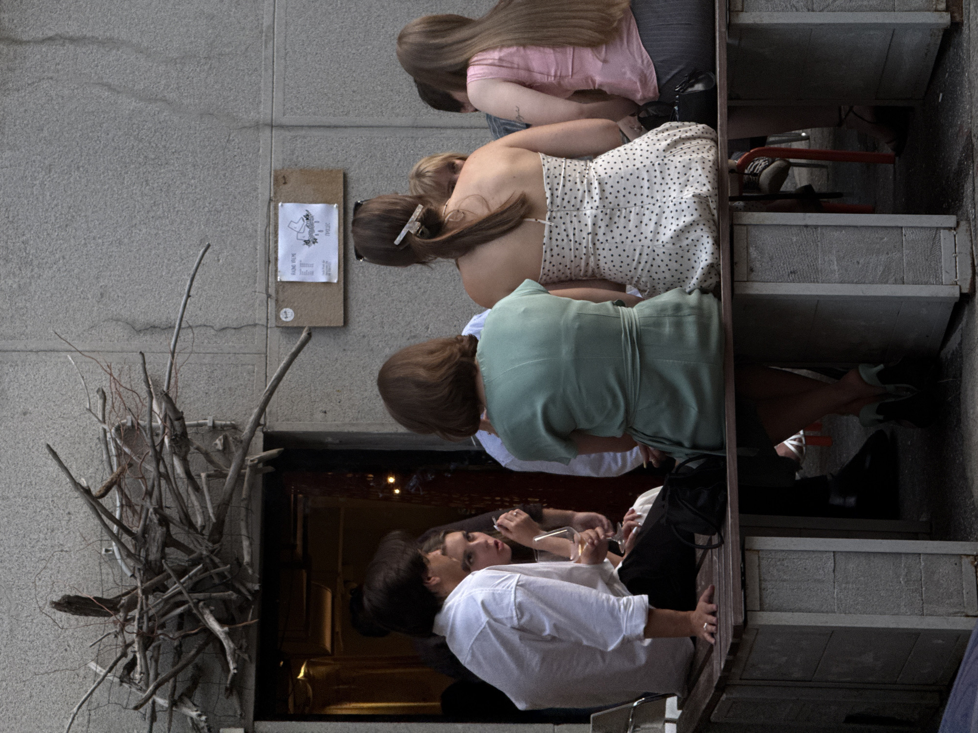 A group of people are sitting on a bench outside what appears to be a building with a concrete wall. In the foreground, a man in a white shirt and dark pants is seated on the left, with his arm resting on the bench. Next to him is a woman wearing a white dress with black polka dots. To her right, another woman in a light green dress is also seated. Further to the right, a woman in a pink top and dark skirt is visible. The background shows a doorway with warm lighting and a sign on the wall above the bench. There is also a decorative arrangement of branches above the doorway. The scene suggests an outdoor seating area, possibly a cafe or restaurant, in Belgrade, Serbia, given the context of the location. The people appear to be engaged in conversation or simply enjoying the outdoor ambiance.