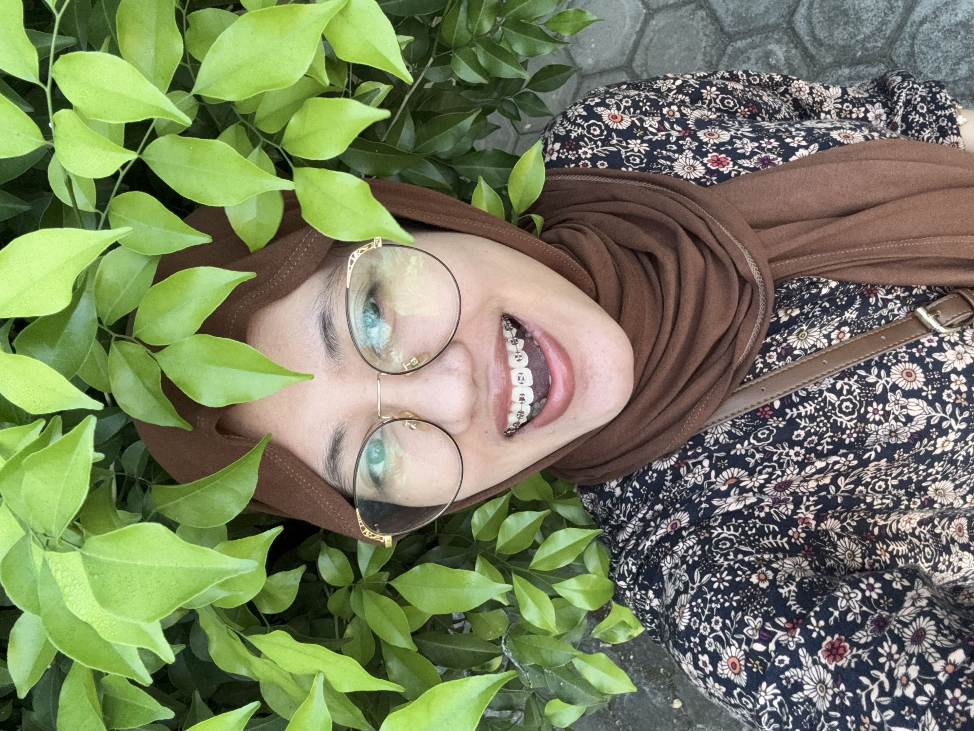 A woman in a brown hijab, eyeglasses, and a floral-patterned shirt smiles at the camera, her braces visible. She is positioned in front of a backdrop of vibrant green leaves, suggesting an outdoor setting. The background also shows a paved surface, possibly a sidewalk or pathway, consistent with an urban environment.