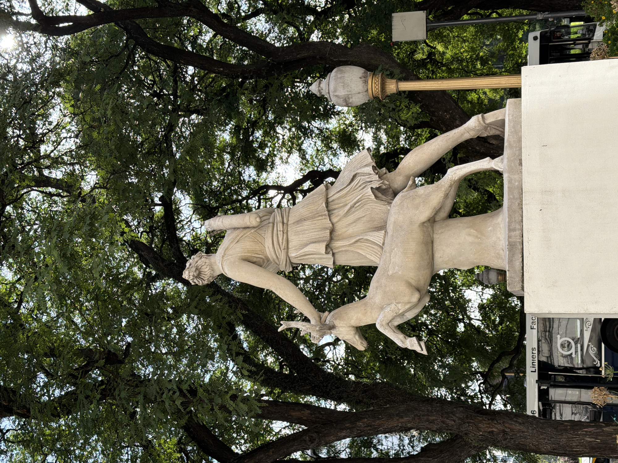 The image shows the "Diana Cazadora" statue in the city of Buenos Aires, Argentina. The statue depicts Diana, the Roman goddess of the hunt, with a deer. The statue is made of stone and stands on a white rectangular base. Behind the statue are tall trees with green leaves. In the background there is a truck and a street light.